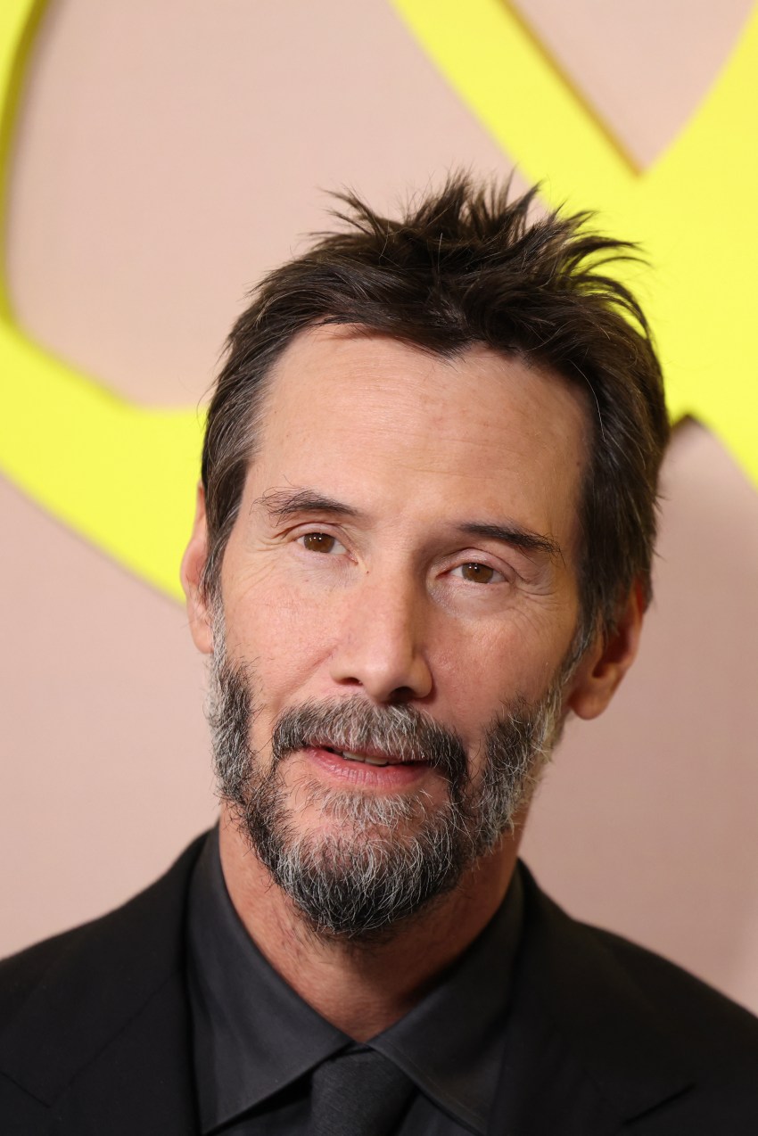 Keanu Reeves with dark spiky hair and a short beard wearing an all-black suit at a premiere event with a yellow logo backdrop.