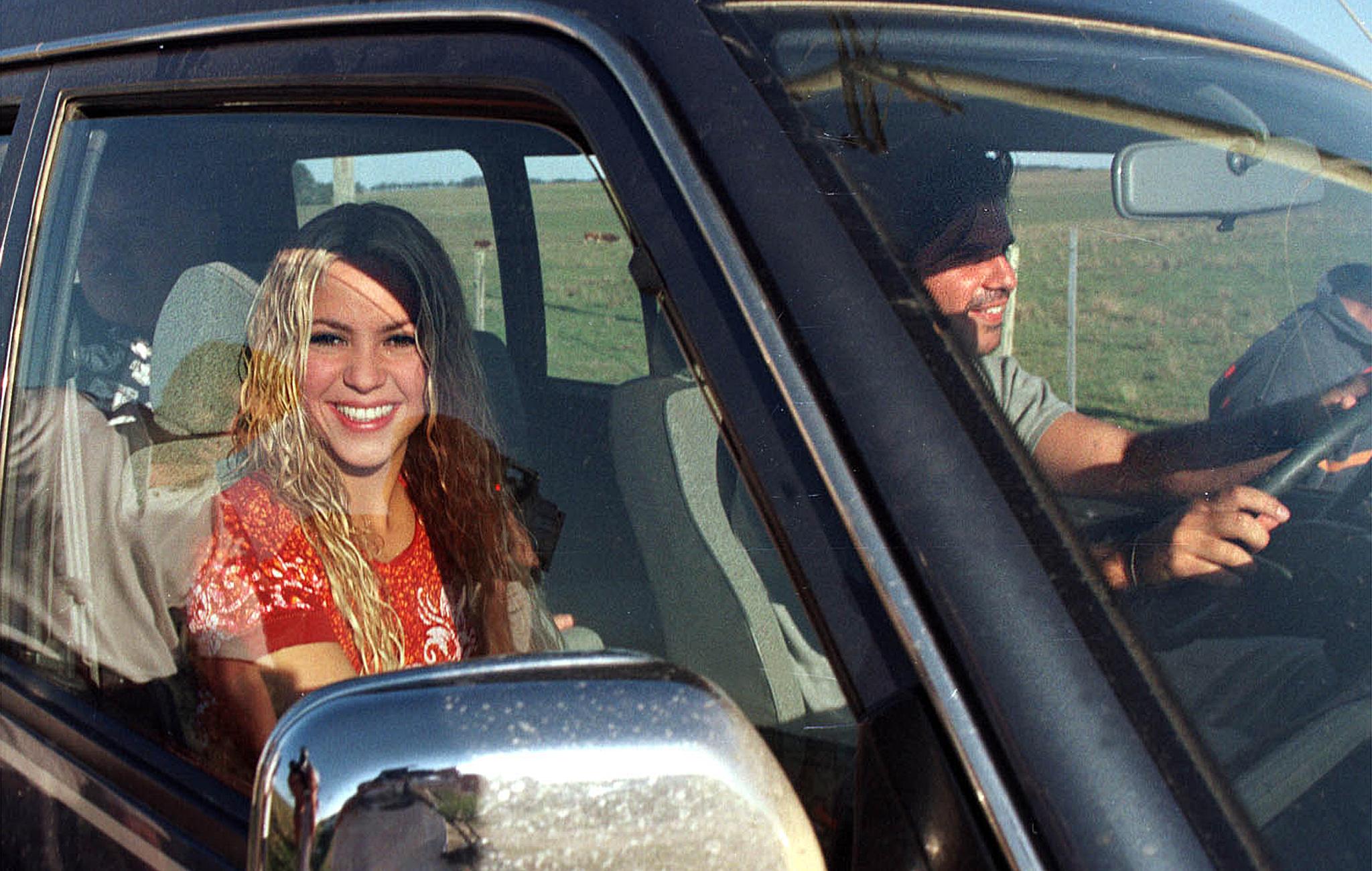 A smiling young woman with long wavy hair sits in the passenger seat of a truck beside a man at the steering wheel, green fields visible through the windows.