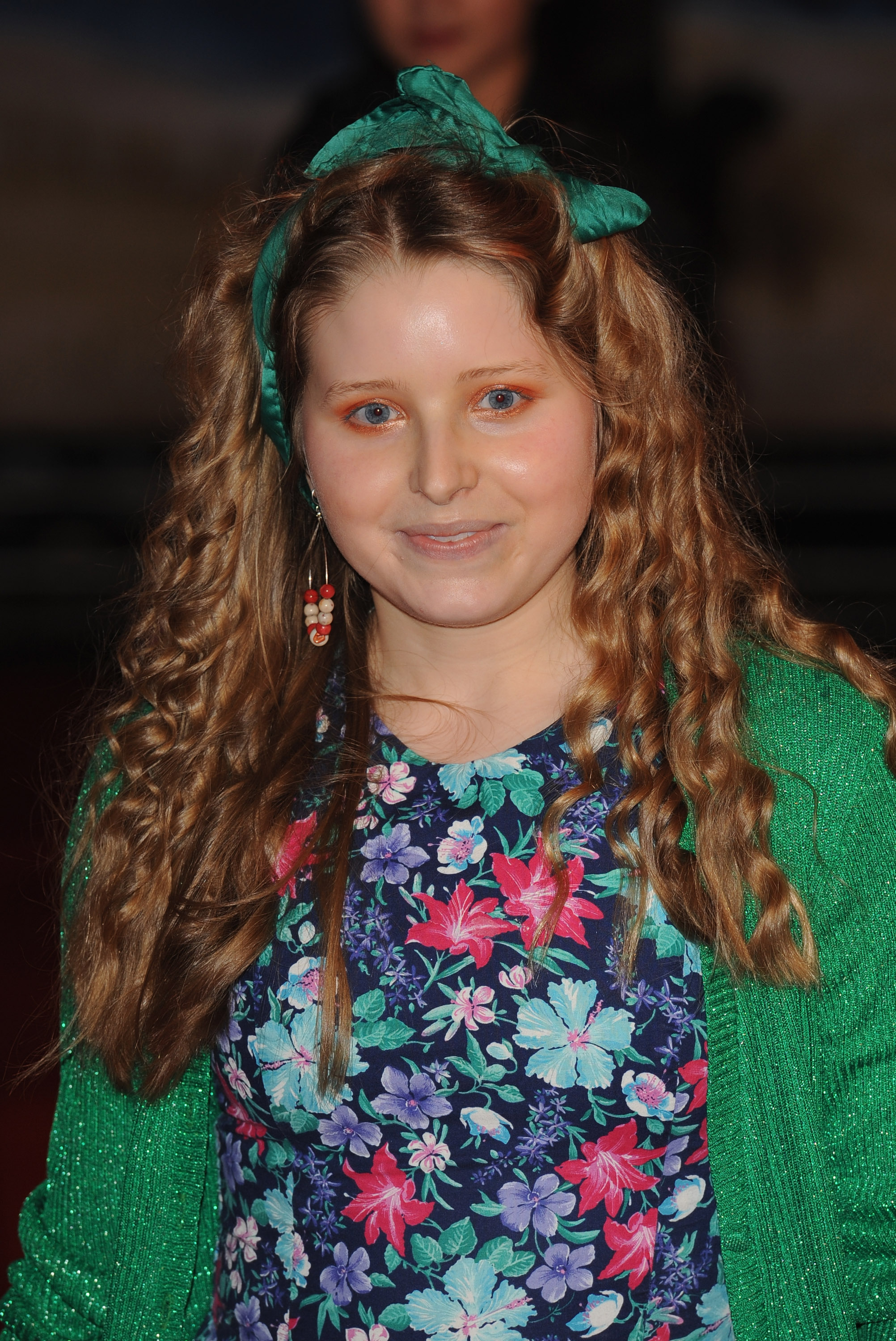 Young woman with curly auburn hair and a green bow, wearing a floral dress and green cardigan on a red carpet.