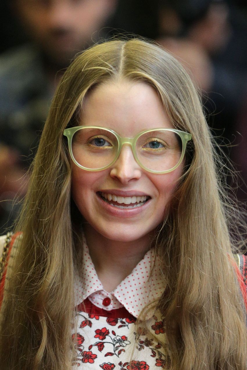 Jessie Cave smiling, wearing round green glasses and a floral top with long blonde hair at a film premiere.