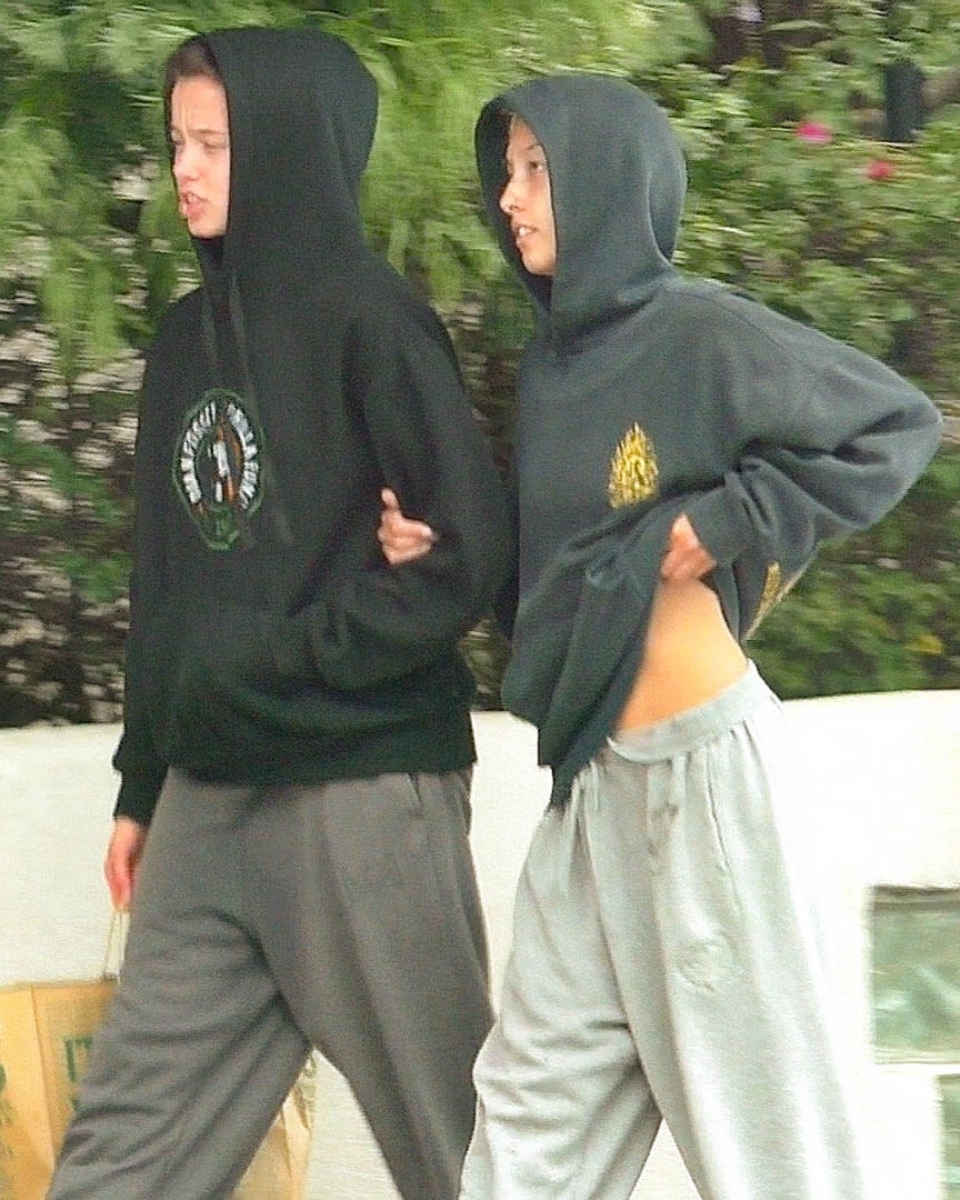 Two people in dark oversized hoodies and grey sweatpants walking side by side outdoors near greenery.