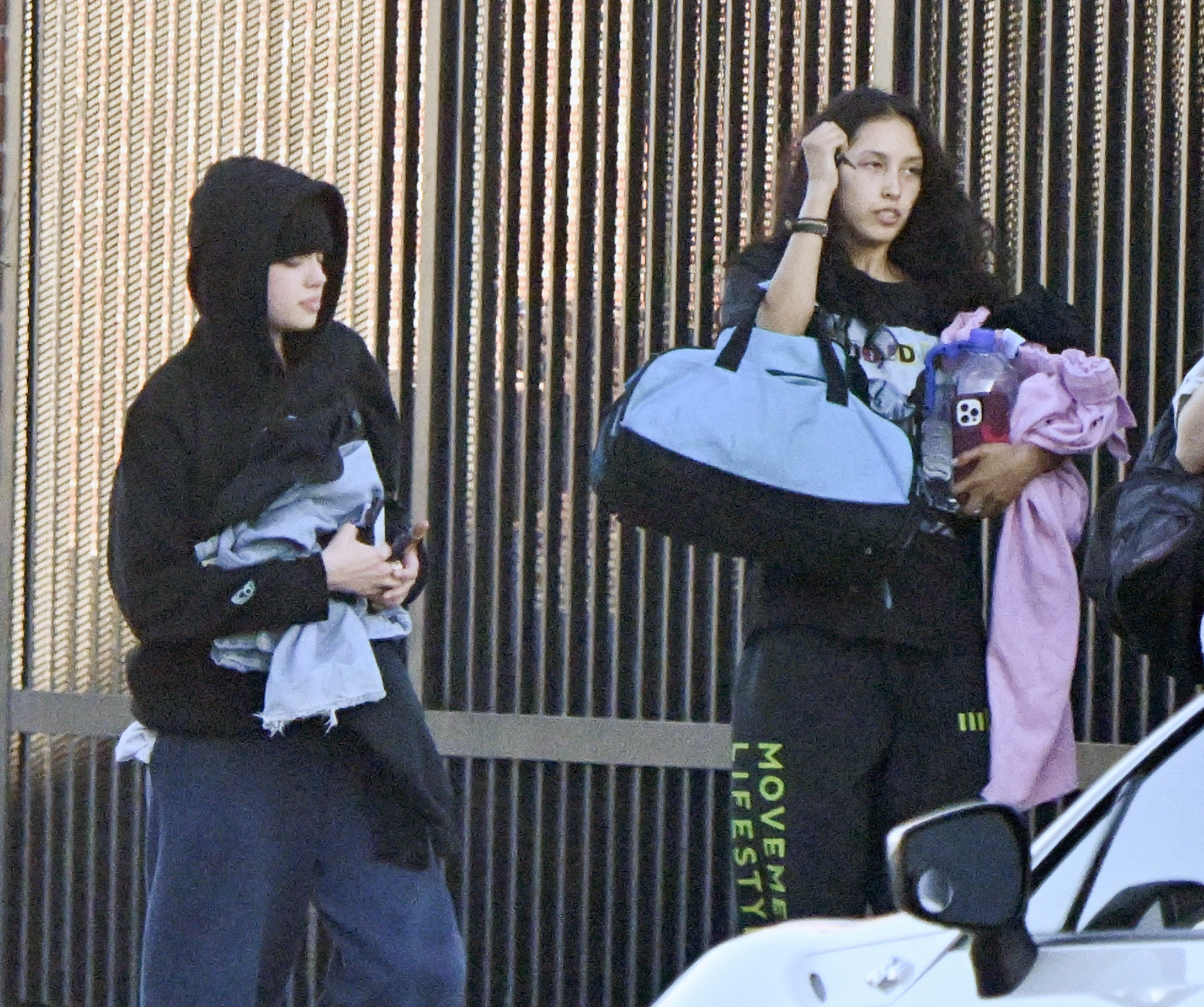 Two young women in black hoodies outside a studio, one carrying a blue duffel bag and pink clothing, the other holding a light blue garment.