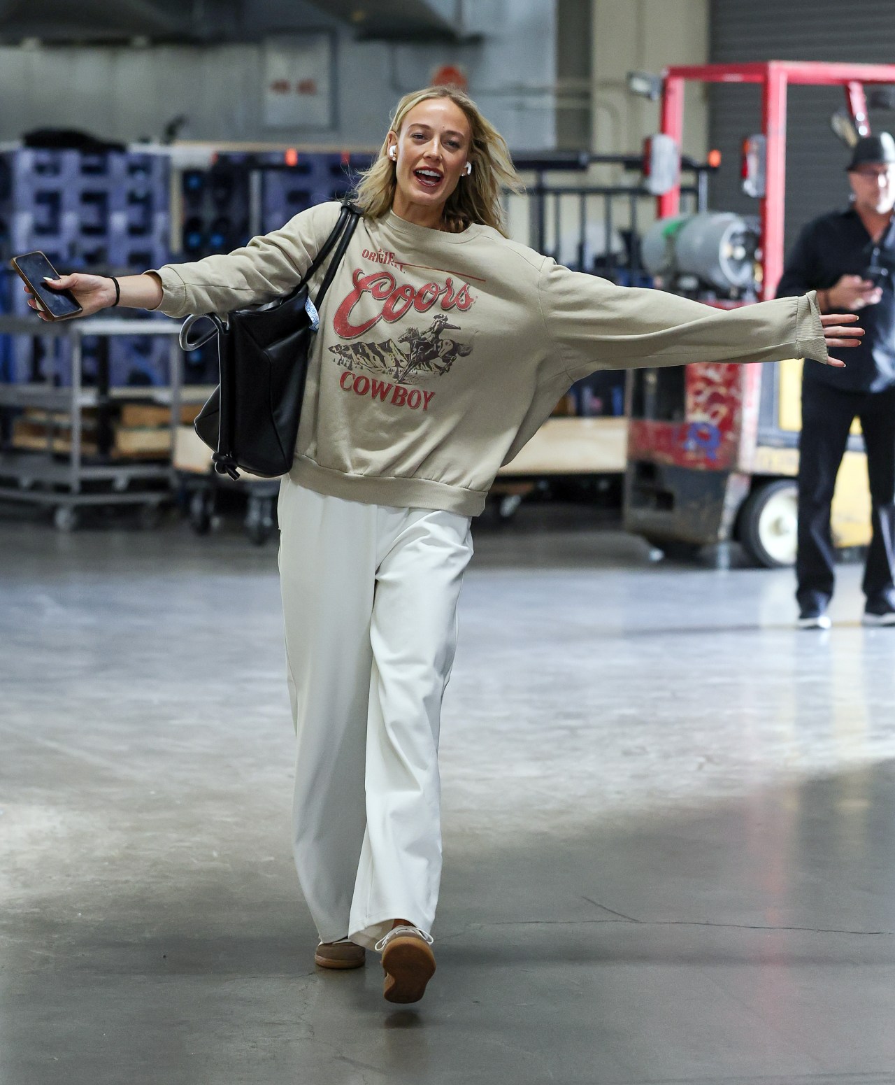 Blonde woman in a Coors Cowboy sweatshirt and white pants walks with arms outstretched, smiling, inside an arena loading area.