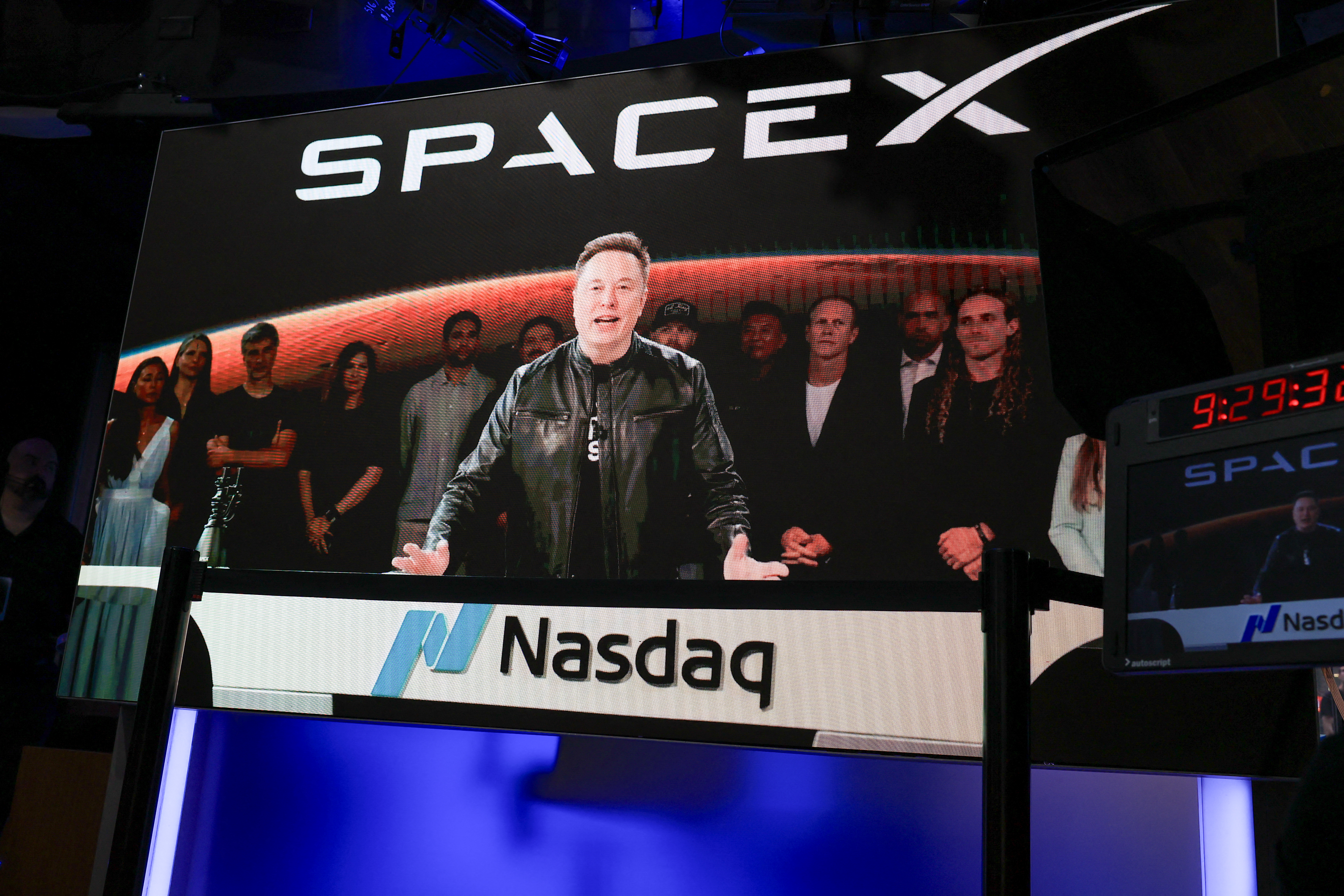 Large Nasdaq screen displaying SpaceX branding and a video of Elon Musk speaking, surrounded by a group of people, at the Nasdaq MarketSite in New York.