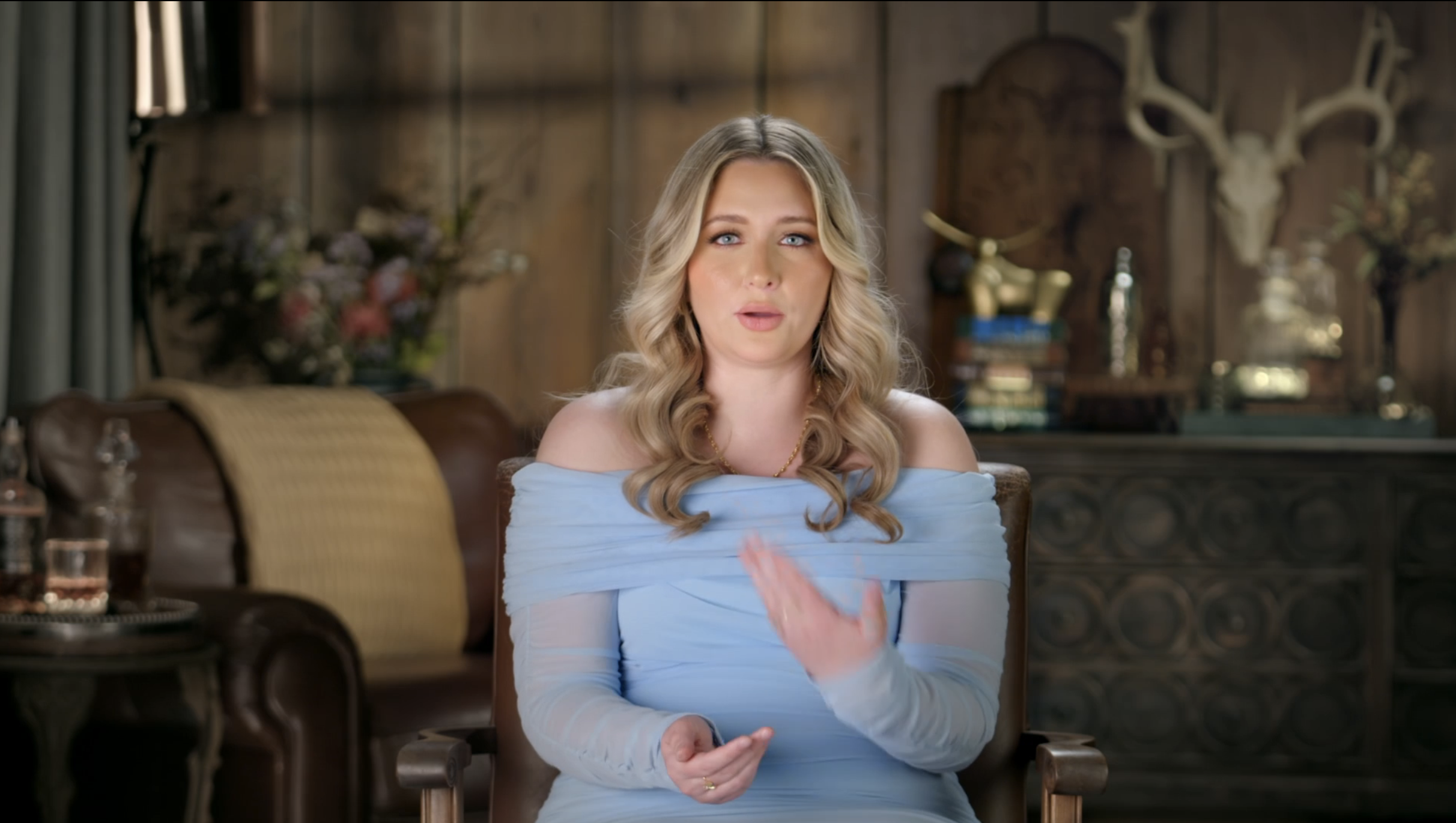Blonde woman in a light blue off-the-shoulder dress seated in a carved wooden chair, gesturing with one hand during an interview in a rustic room.