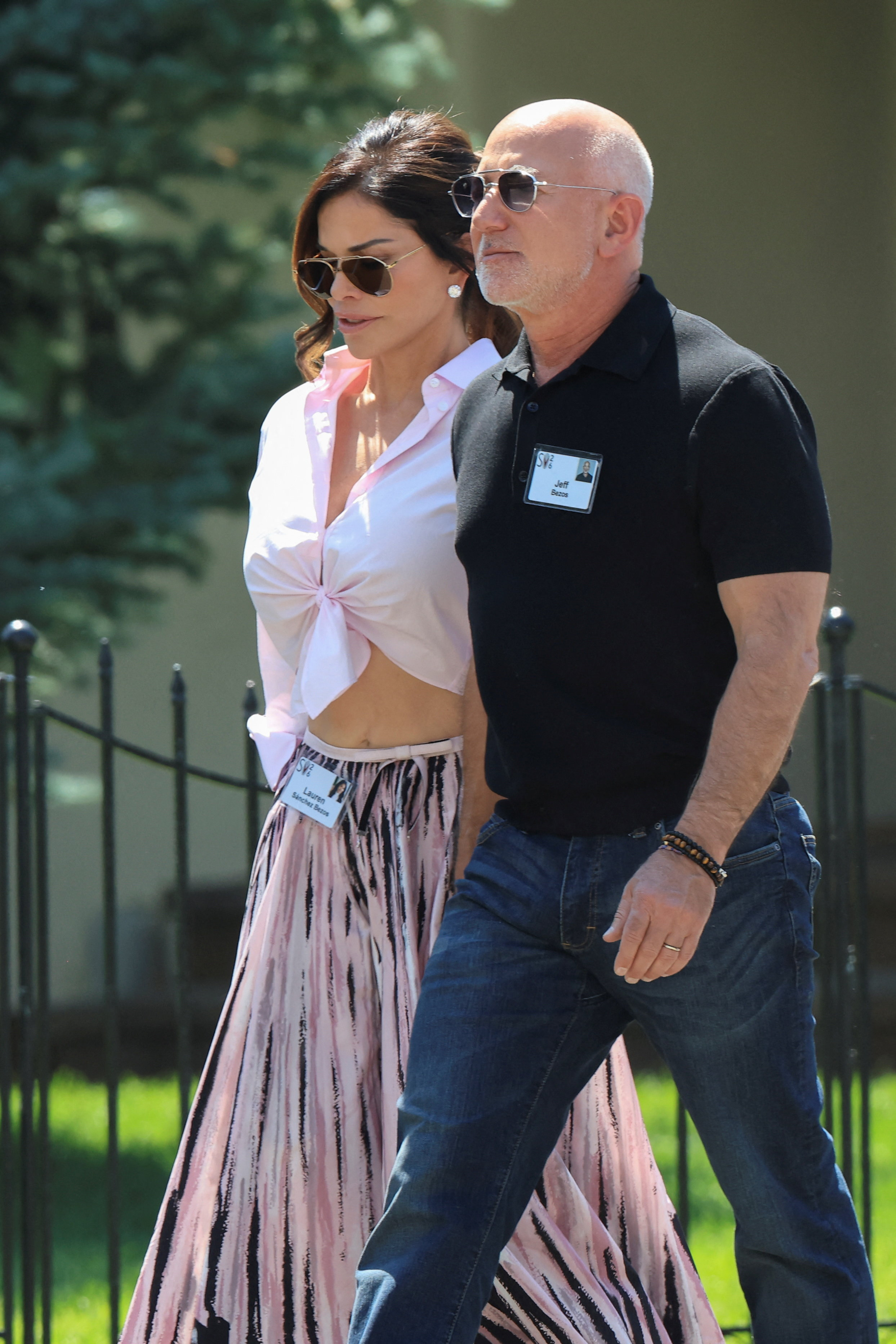 Jeff Bezos in a black polo shirt and Lauren Sanchez in a pink tied shirt and printed skirt walk together outdoors at Sun Valley Resort.