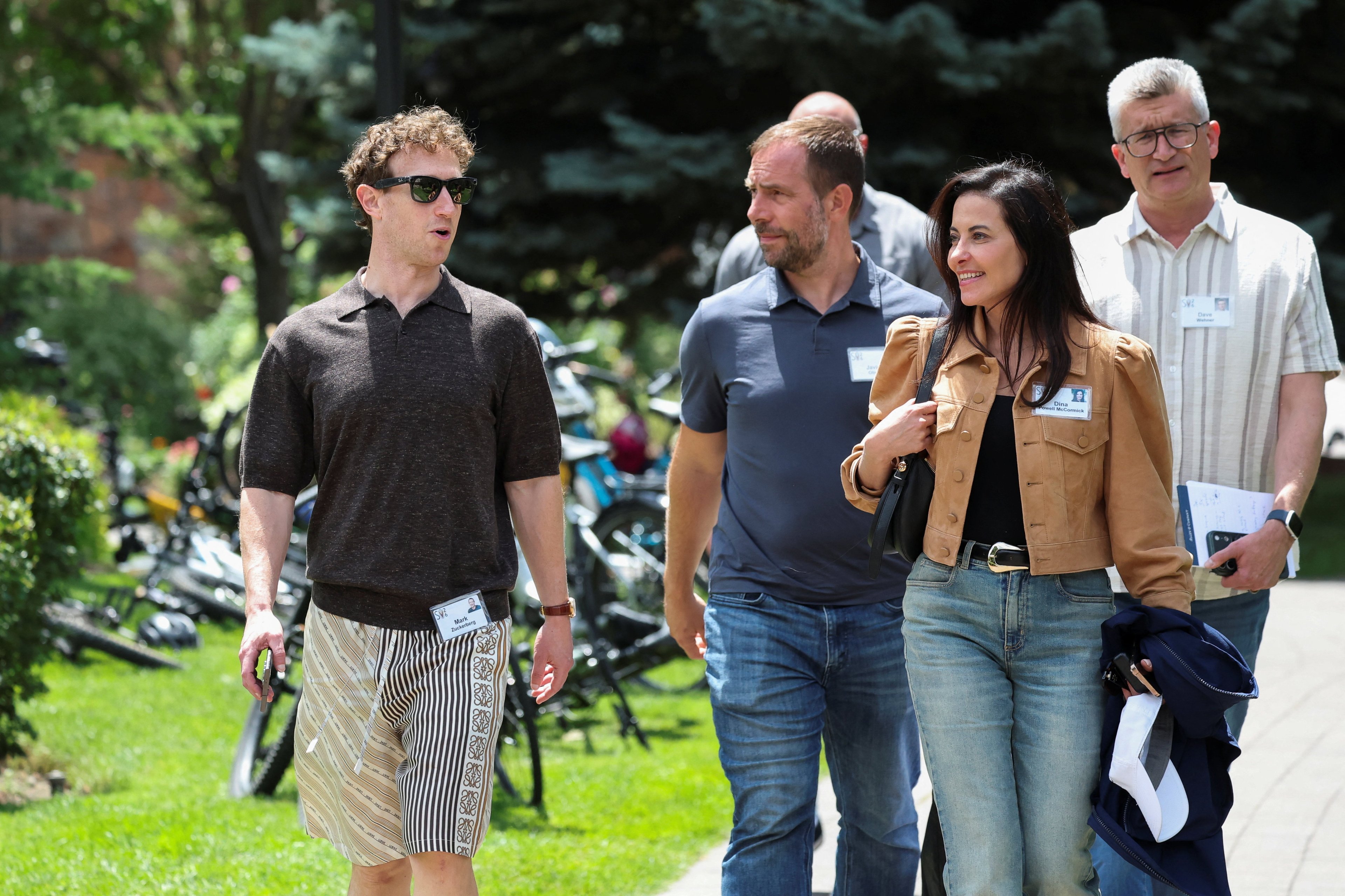 Mark Zuckerberg in sunglasses and shorts walks with Dina Powell McCormick and Meta executives on a sunny outdoor path in Sun Valley, Idaho.
