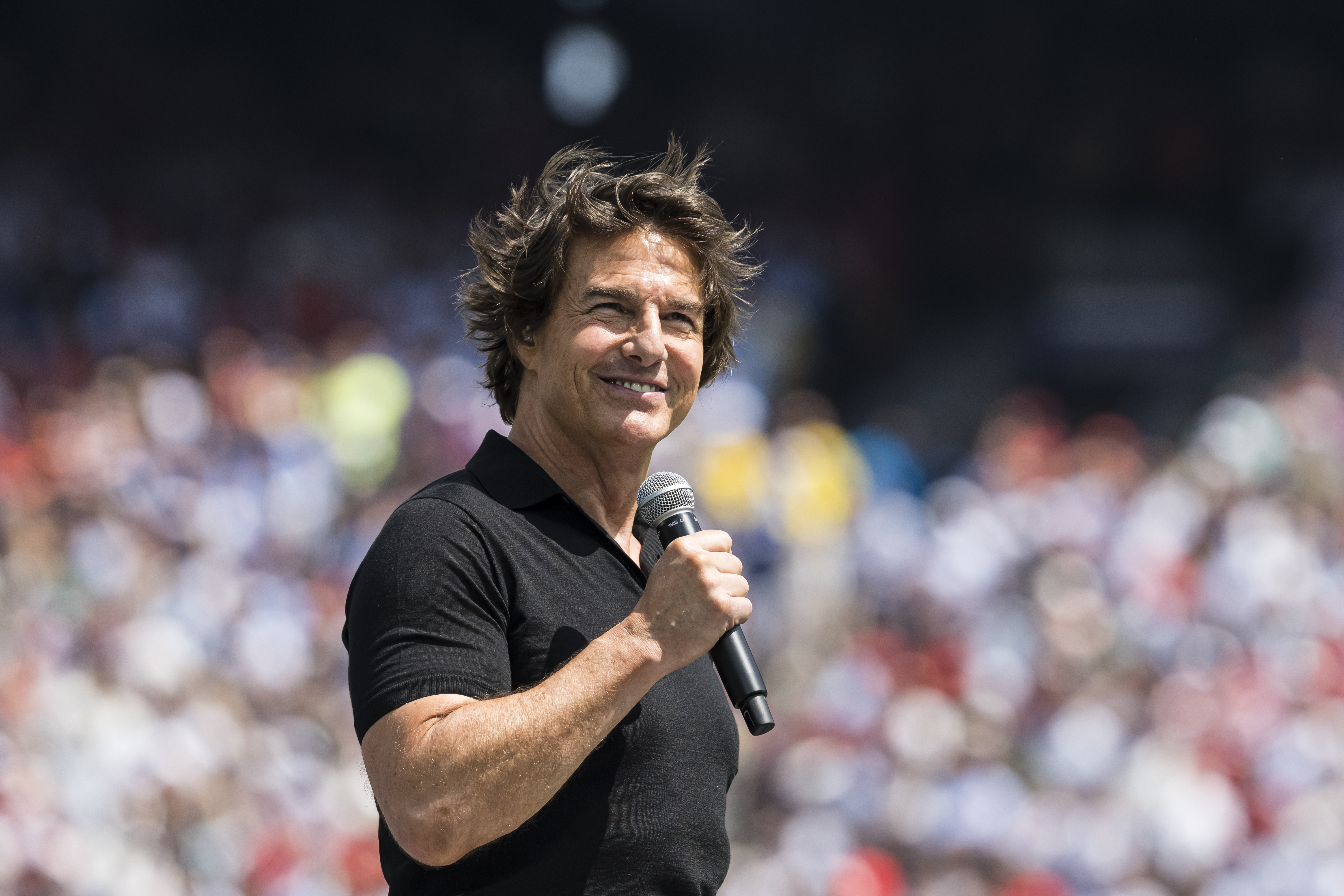 Man in black polo shirt holding a microphone and smiling before a blurred stadium crowd outdoors.