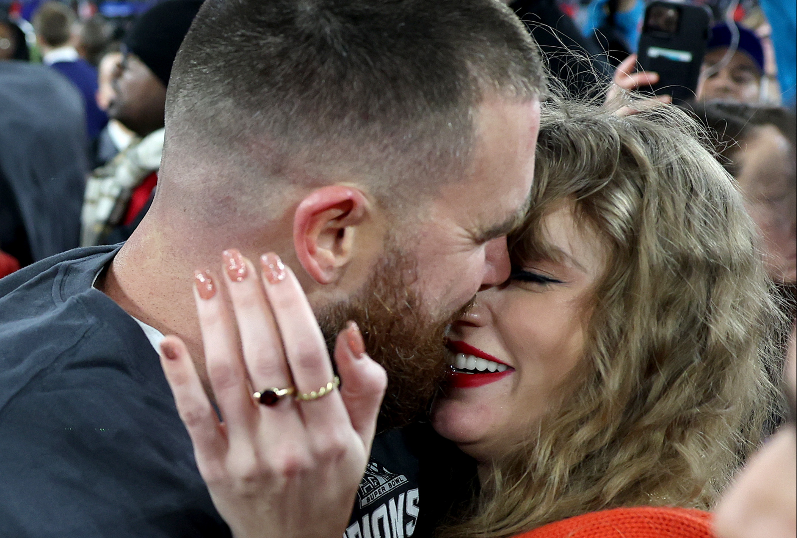 Travis Kelce and Taylor Swift embracing and smiling on the football field after the AFC Championship game, Swift wearing a red sweater.