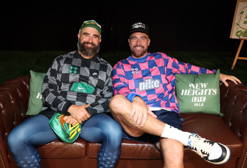 Jason Kelce and Travis Kelce seated on a brown leather couch wearing matching Nike checkered jerseys at New Heights Live in LA.