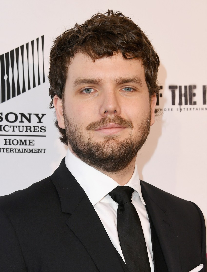 Young man with curly brown hair and beard in a black suit and tie at a Sony Pictures event.