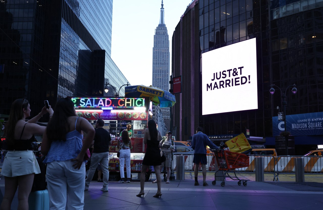 Crowd of people on a New York City sidewalk near Madison Square Garden looking at a bright billboard reading "JUST&T MARRIED!" with the Empire State Building in the distance.