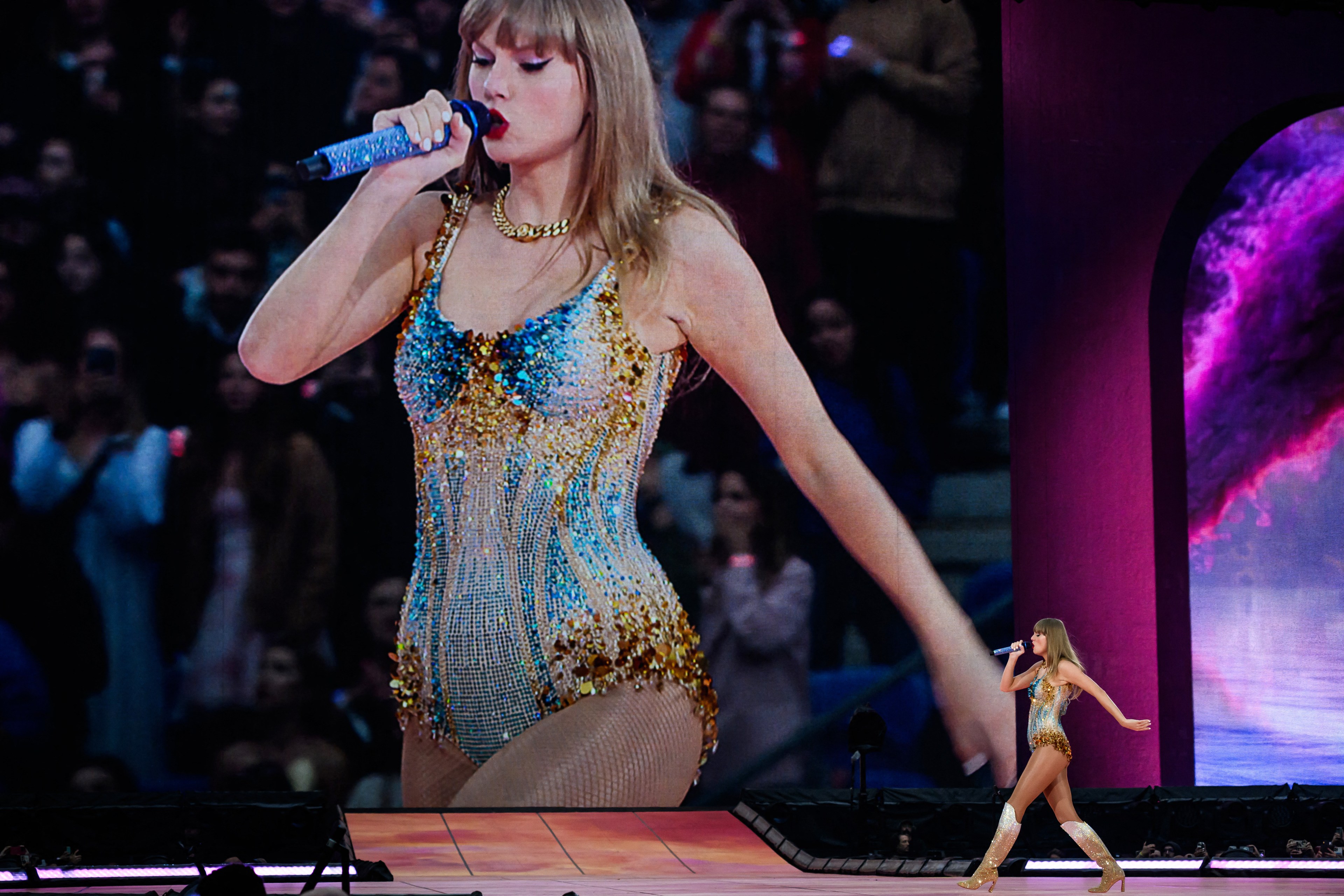 Taylor Swift performing on stage in a sequined bodysuit, singing into a microphone, with a massive close-up of her on the LED screen behind her.