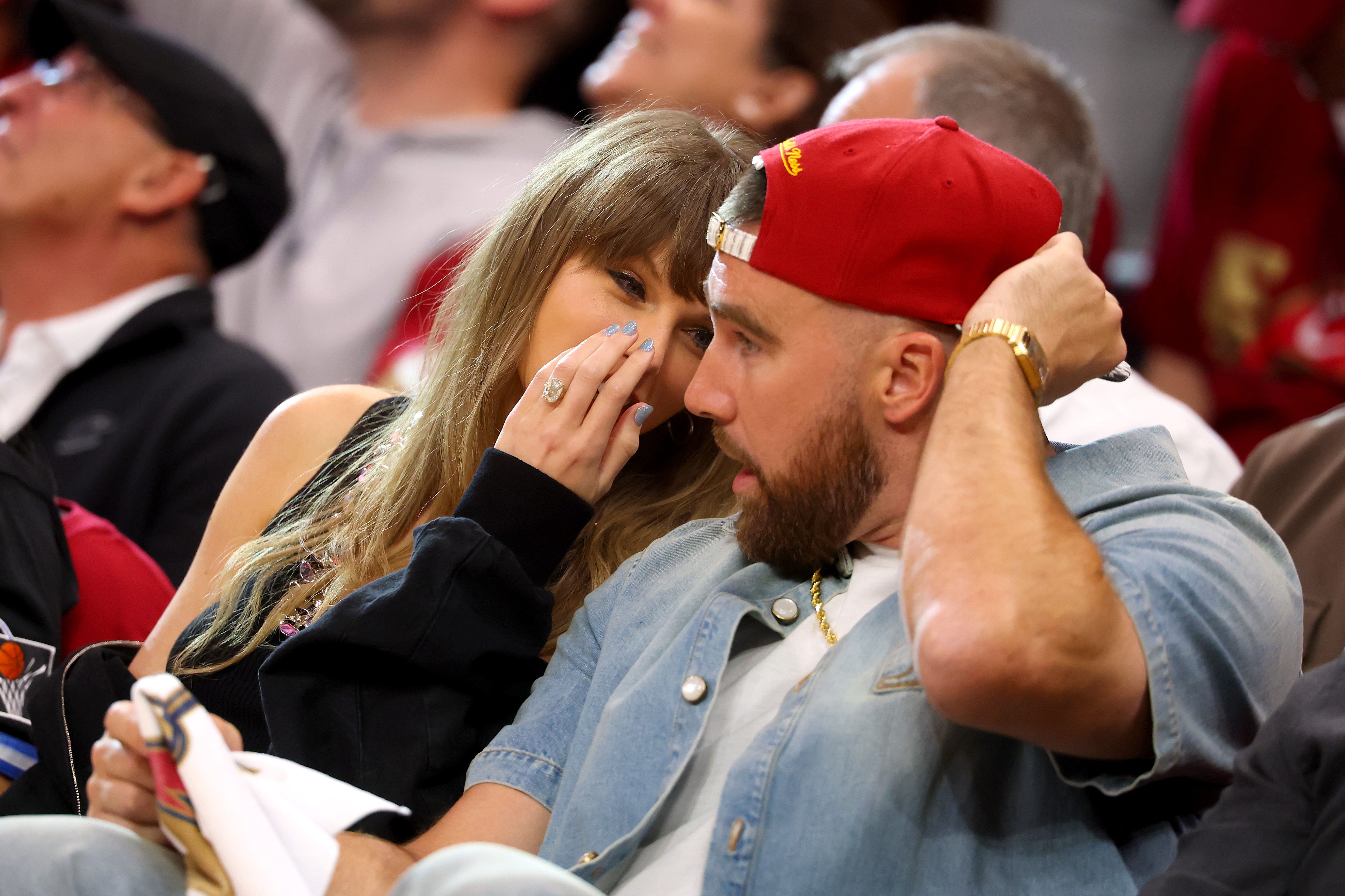 Click to open the image gallery A woman with long wavy hair whispers to a bearded man in a red cap and denim jacket while seated in a crowded basketball arena.