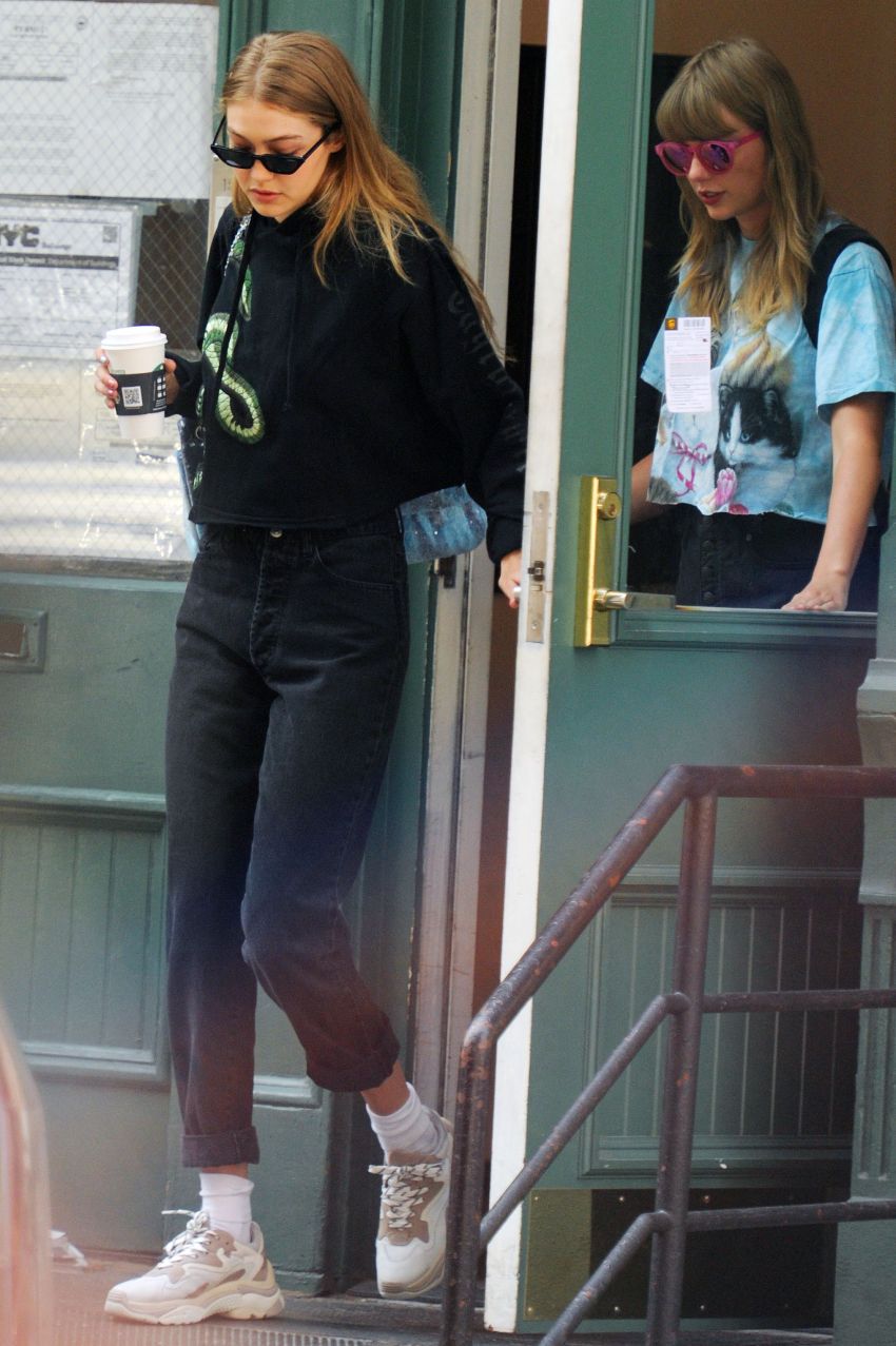 A woman in a black snake-print hoodie, dark jeans, and white sneakers carries a coffee cup while exiting a building, with another woman in a blue cat tee and pink sunglasses visible in the doorway.