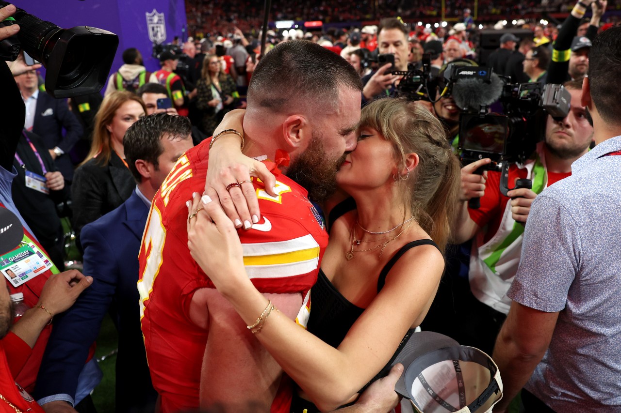 Travis Kelce in a red Kansas City Chiefs jersey kisses Taylor Swift on the crowded field after Super Bowl LVIII at Allegiant Stadium.