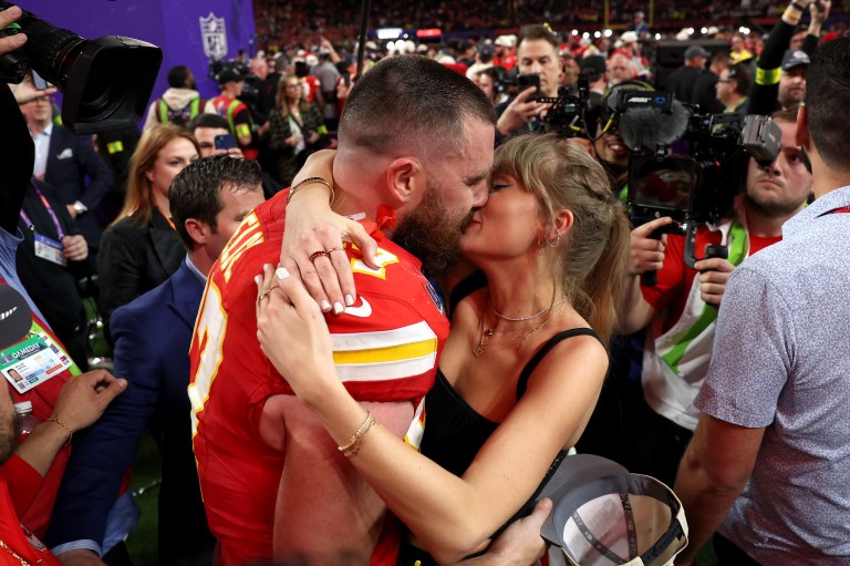 Travis Kelce in a red Kansas City Chiefs jersey kisses Taylor Swift on the crowded field after Super Bowl LVIII at Allegiant Stadium.