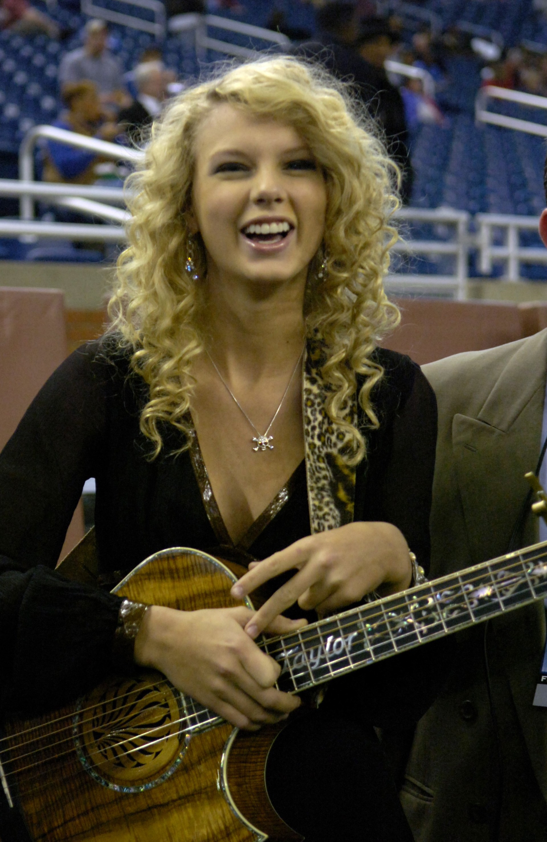 A young blonde curly-haired woman smiling and holding a Taylor-brand acoustic guitar inside a football stadium.