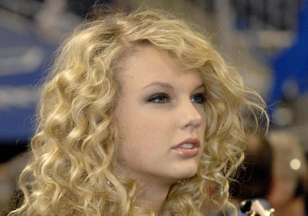 Close-up of a young woman with long curly blonde hair and blue eyes against a blurred blue background.