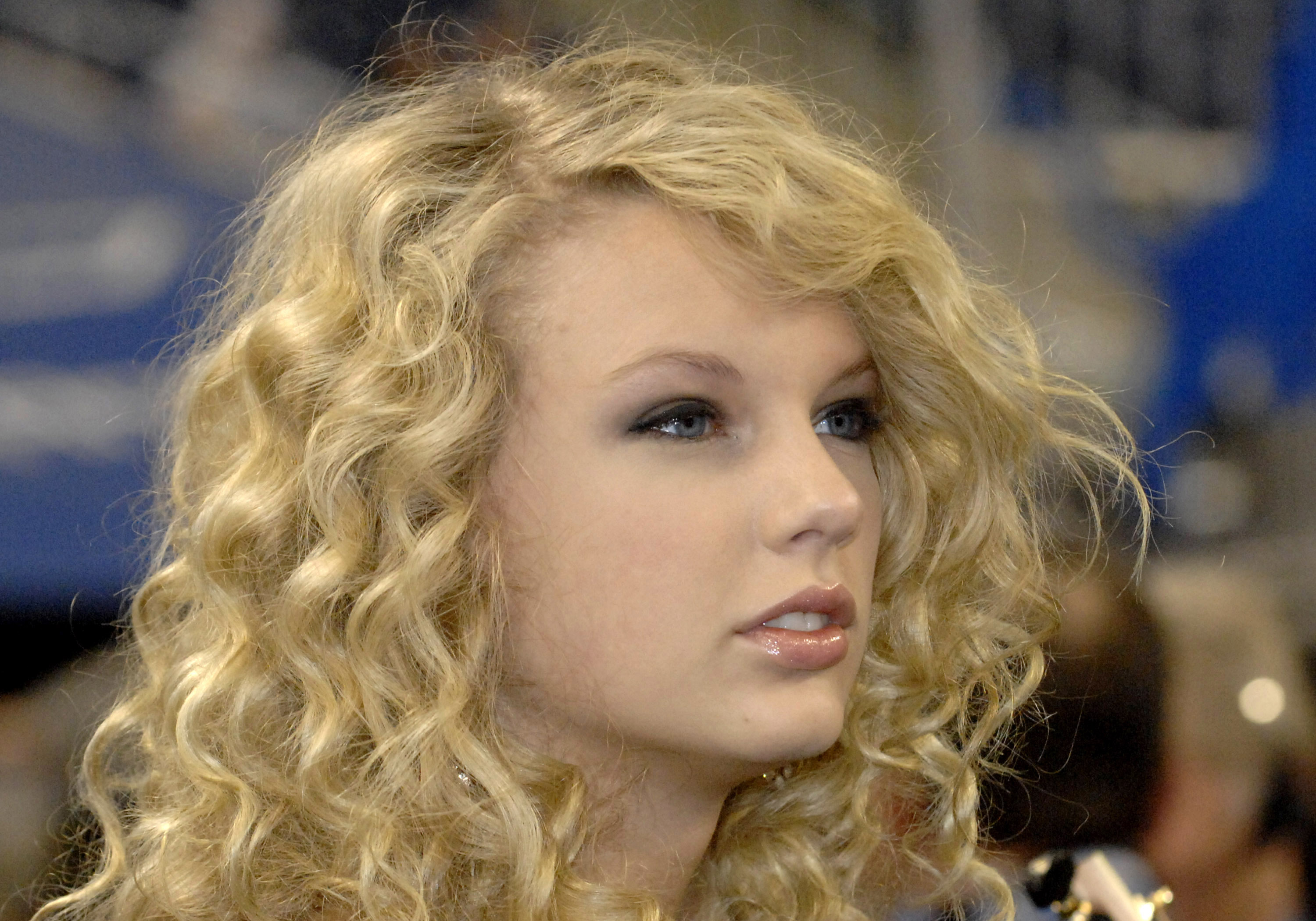 Close-up of a young woman with long curly blonde hair and blue eyes against a blurred blue background.