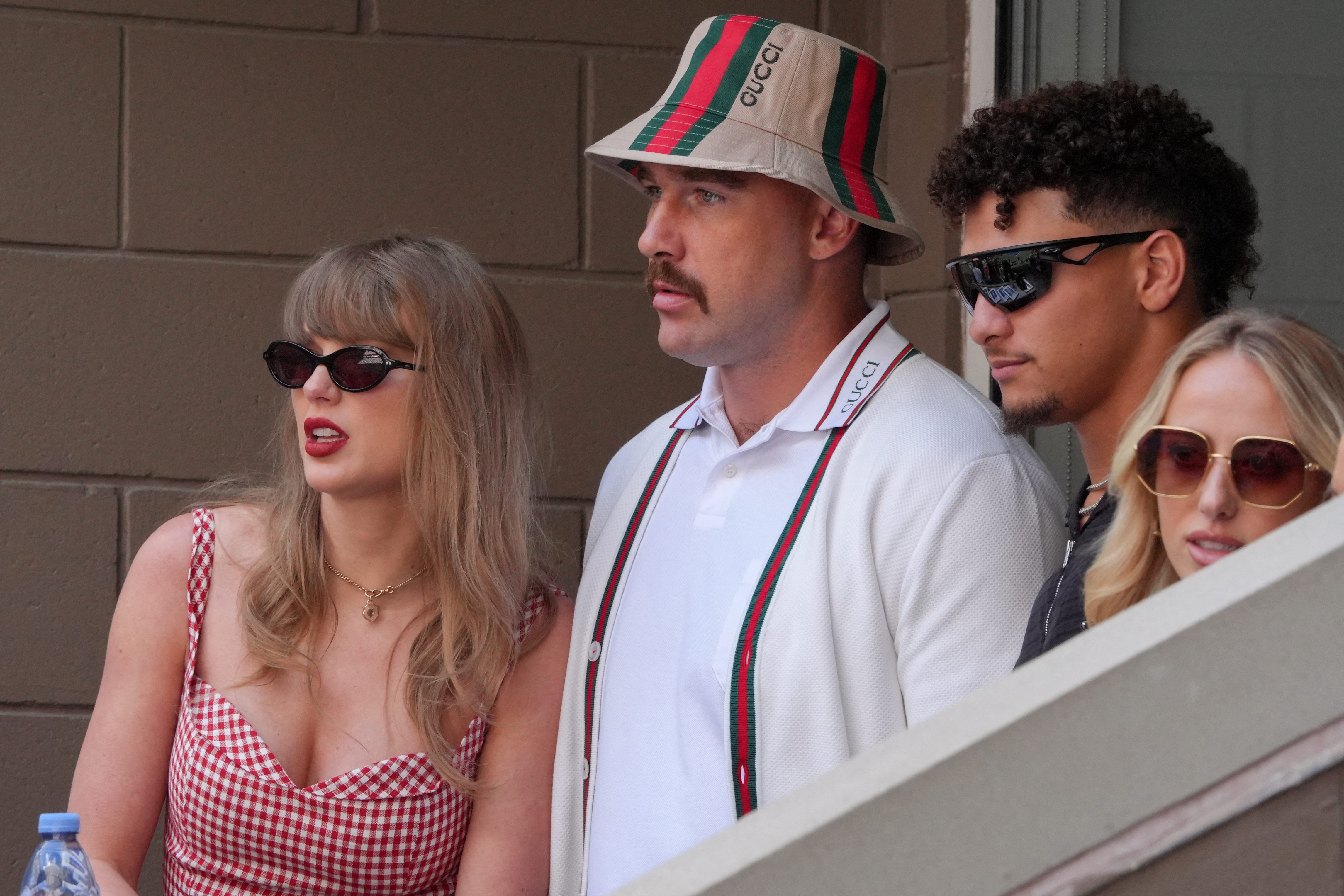 Four people stand in a stadium: a woman in a red gingham dress, a man in a Gucci bucket hat, a curly-haired man in sunglasses, and a blonde woman in sunglasses.