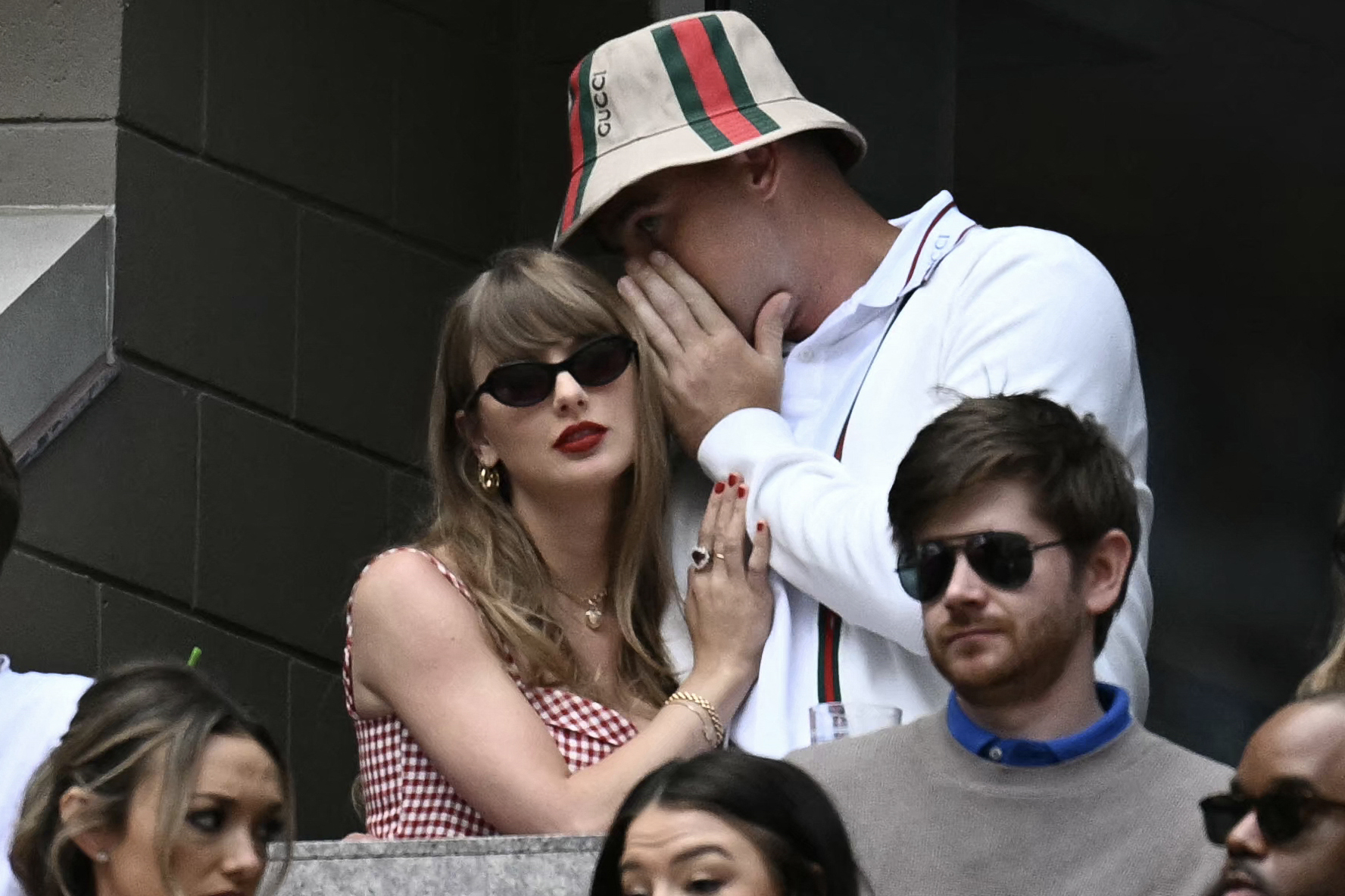 Man in Gucci bucket hat cups his hand to whisper into the ear of a woman in a red gingham dress and dark sunglasses at a tennis match.