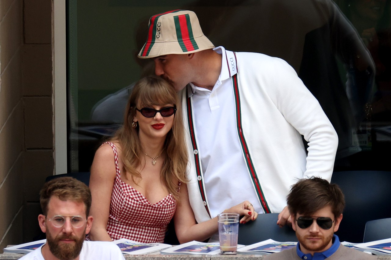 A man in a Gucci bucket hat and white cardigan kisses a blonde woman in a red gingham dress on the head at a tennis stadium.