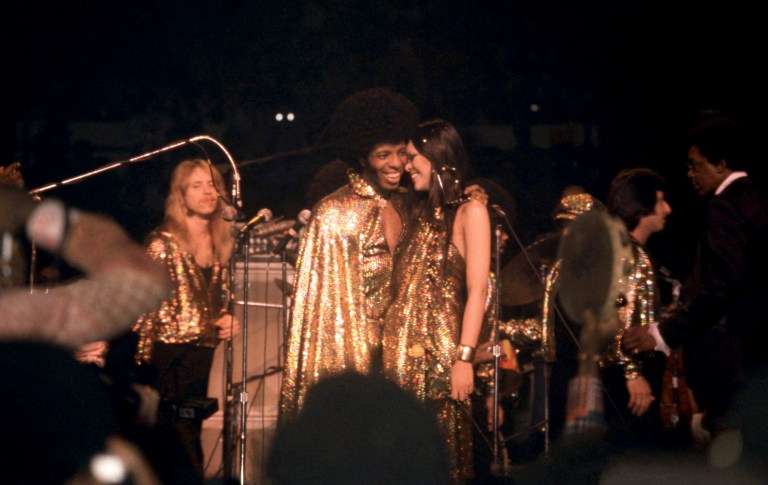 Sly Stone and Kathy Silva in matching gold sequined outfits smiling face-to-face on stage at Madison Square Garden, surrounded by band members.