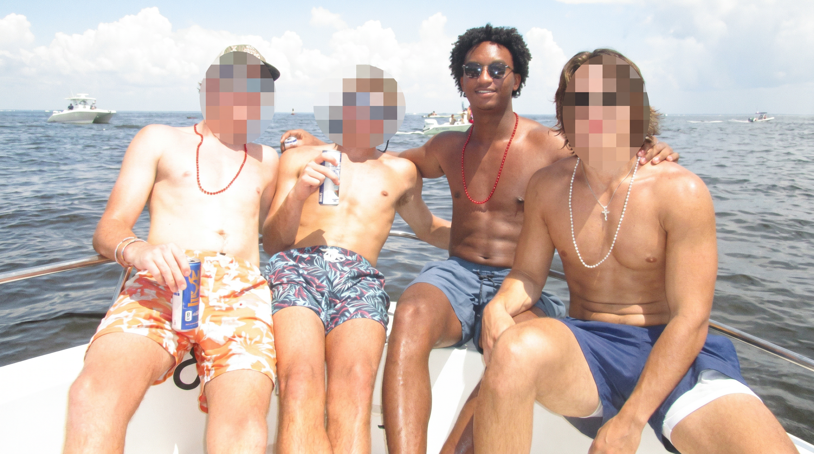 Four shirtless young men in swim trunks sitting on the edge of a boat on open water; three have pixelated faces and one smiles wearing sunglasses and a red beaded necklace.