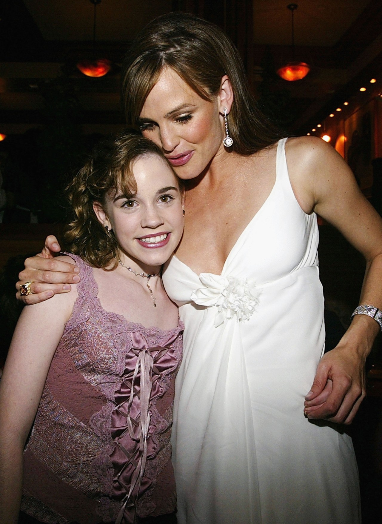 Woman in a white dress hugging a young girl in a pink lace top at an indoor event.