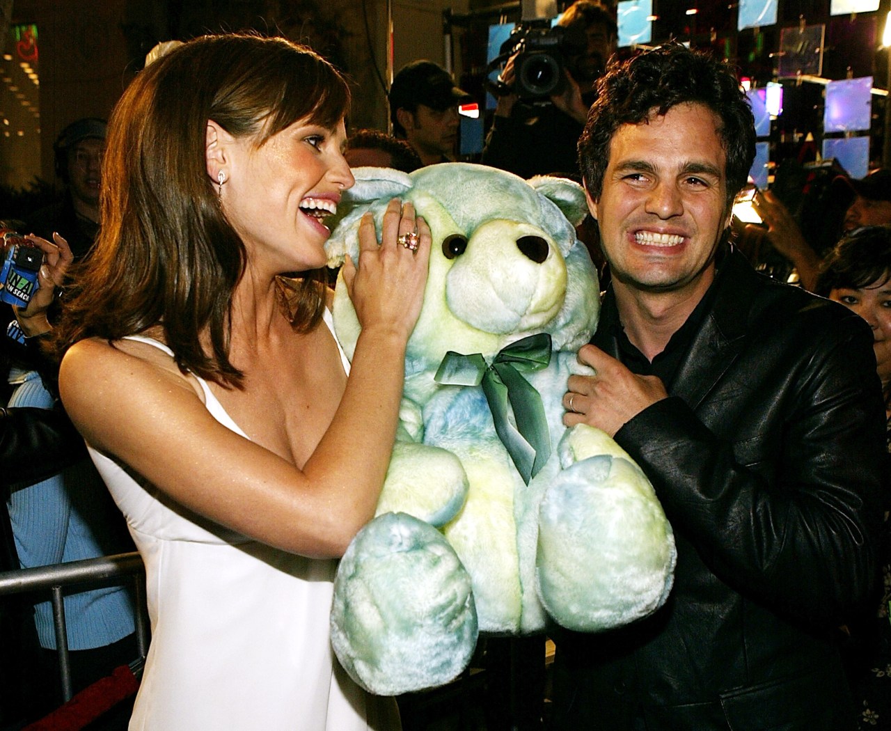 A woman in a white dress and a man in a black leather jacket laughing while holding a large blue and green stuffed teddy bear.