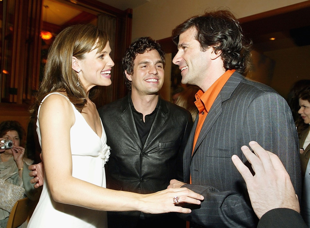 Two men and a woman in a white dress laughing together at an indoor after-party.