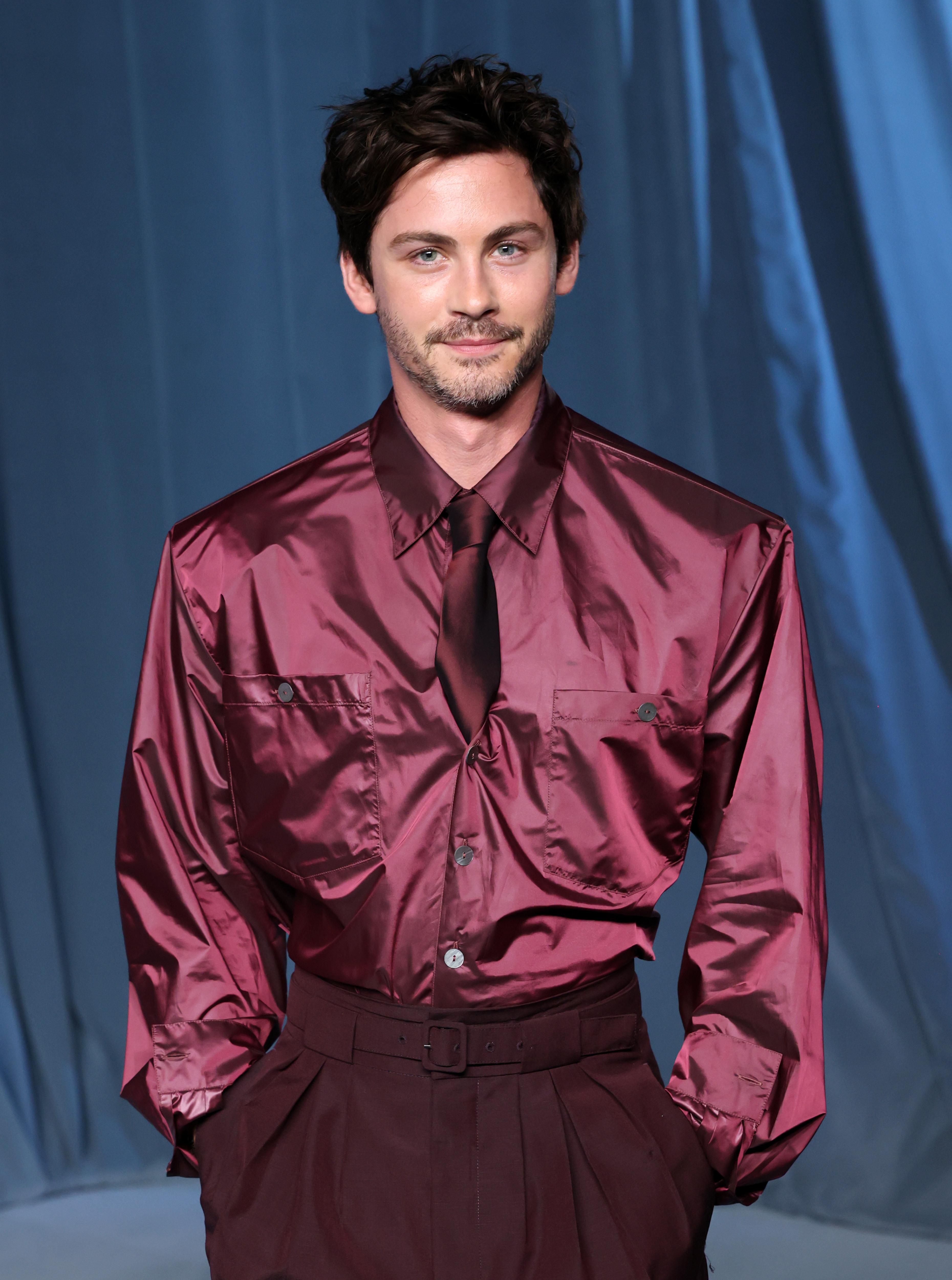 Young man with dark curly hair in a shiny burgundy button-up shirt and dark tie, posing against a blue draped background.
