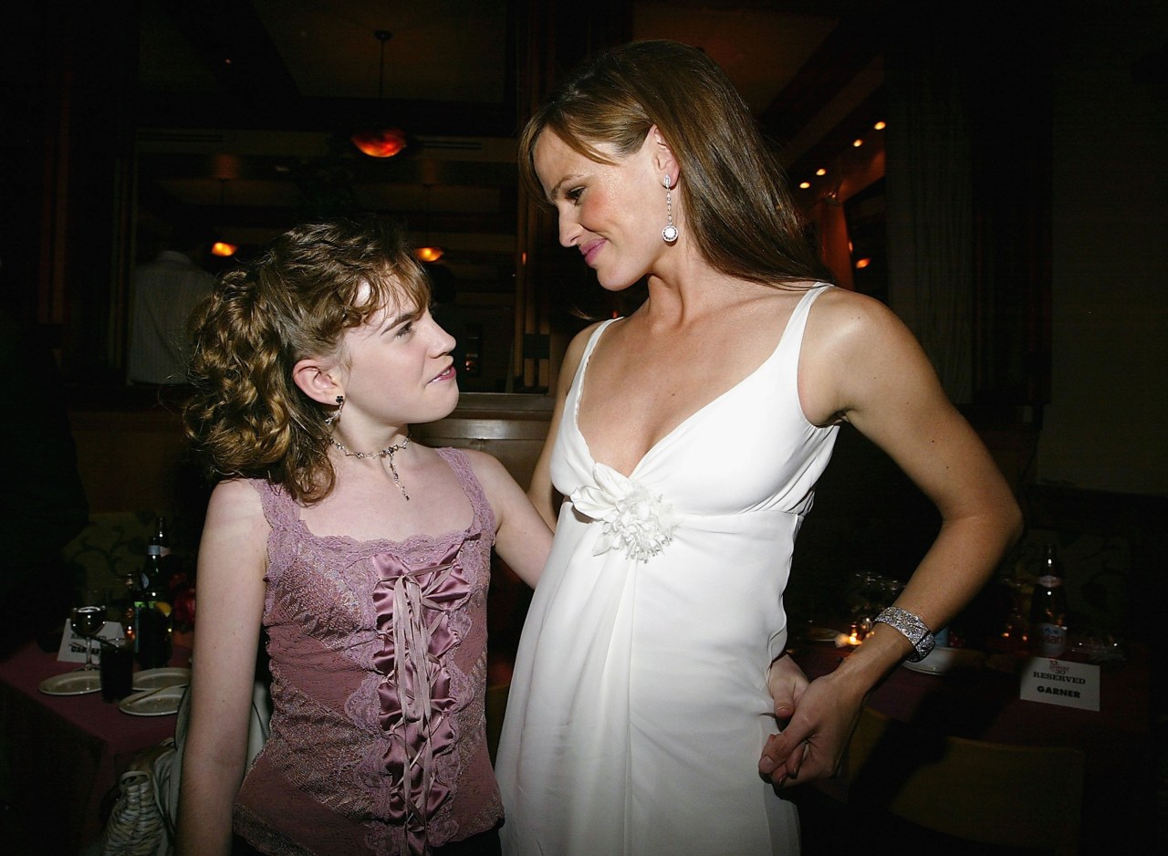 Young girl in a pink lace top talking with a woman in a white dress at a dimly lit after-party.