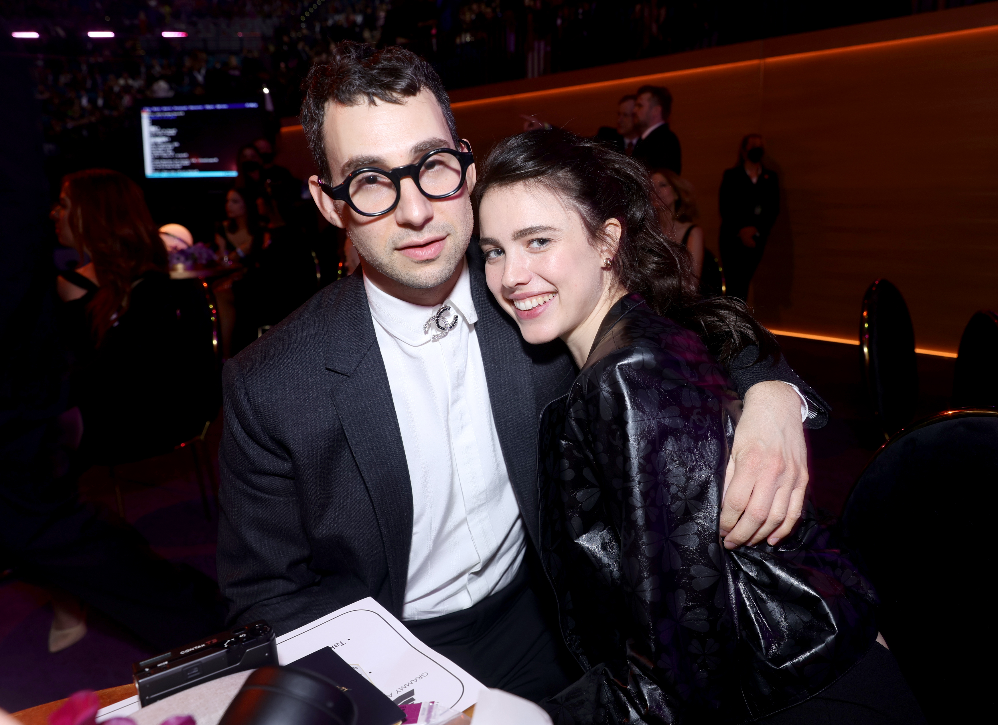 Click to open the image gallery Jack Antonoff in a dark suit with round glasses and Margaret Qualley in a black outfit smile together at the Grammy Awards.