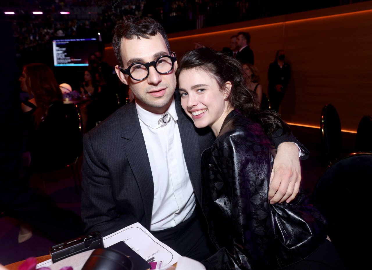 Jack Antonoff in a dark suit with round glasses and Margaret Qualley in a black outfit smile together at the Grammy Awards.