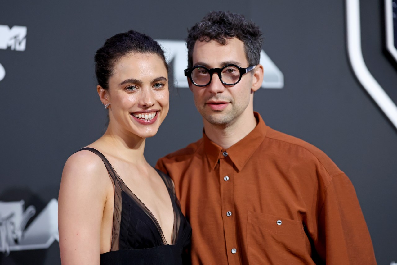 Margaret Qualley in a black dress and Jack Antonoff in an orange shirt and round glasses pose together on the MTV VMAs red carpet.