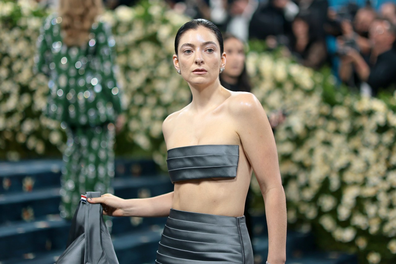 Lorde wearing a gray strapless bandeau top and matching high-waisted skirt on the Met Gala red carpet, surrounded by floral arrangements.
