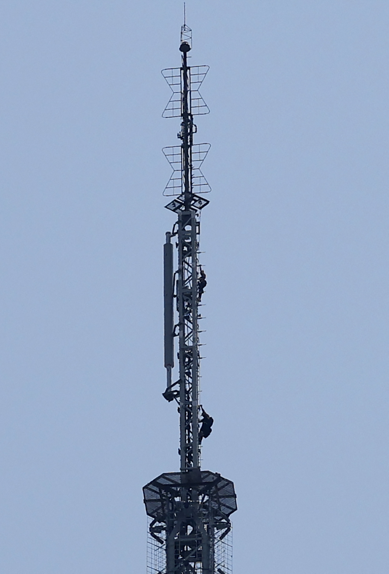 Two people clinging to the spire of the Empire State Building against a pale grey sky, with antenna arrays and tower structure visible.