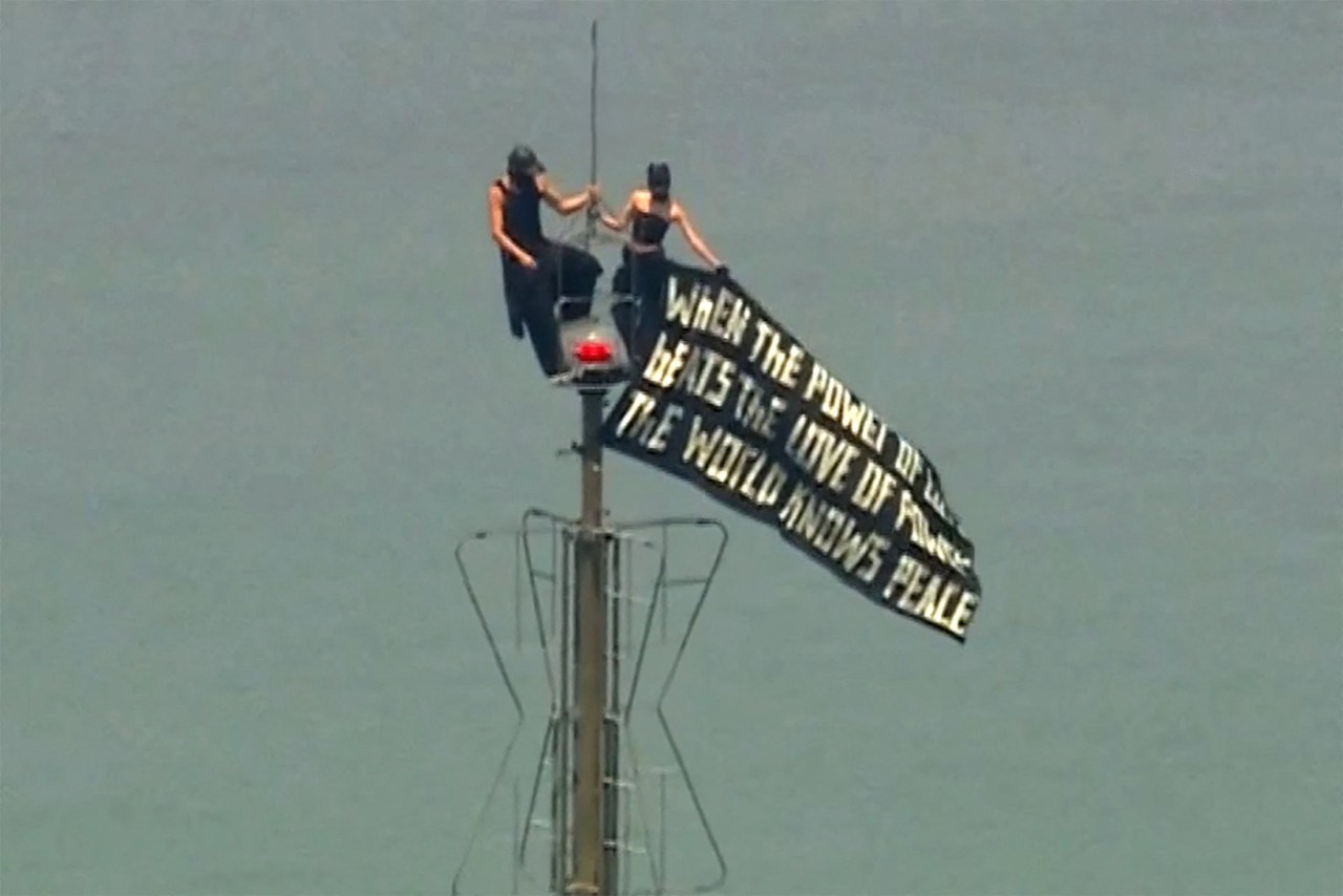 Two people dressed in black stand atop the Empire State Building's antenna mast, holding a large black banner with a peace quote.