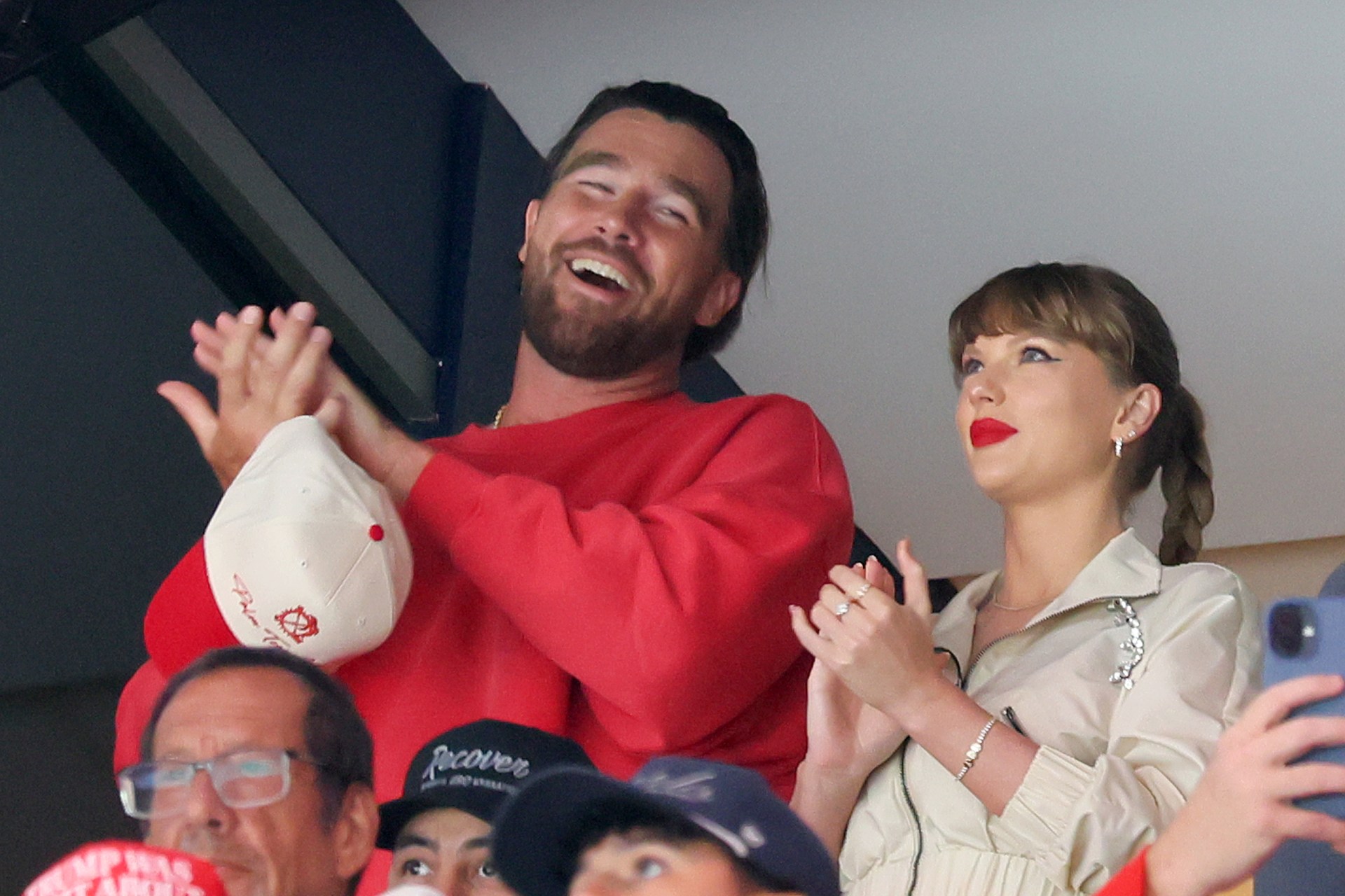 Travis Kelce in a red sweatshirt laughing and clapping next to Taylor Swift, who wears a cream jacket and red lipstick, at a hockey game.