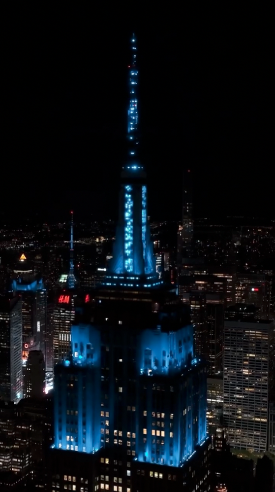 Wide-angle night shot of the Empire State Building illuminated in blue, with the New York City skyline and an H&M sign visible in the background.