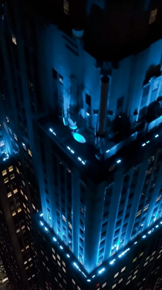 Angled close-up of the Empire State Building's upper floors and terraces lit in blue at night, with city buildings in the background.