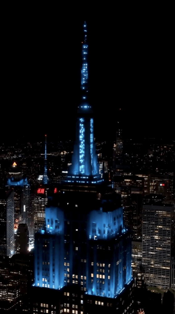 Aerial night view of the Empire State Building lit up in bright blue light, surrounded by the glowing New York City skyline.
