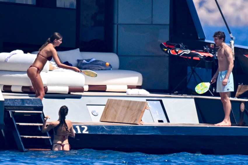 Shirtless young man and woman in bikini playing beach tennis on a yacht deck while another woman in a bikini climbs a ladder from the sea.