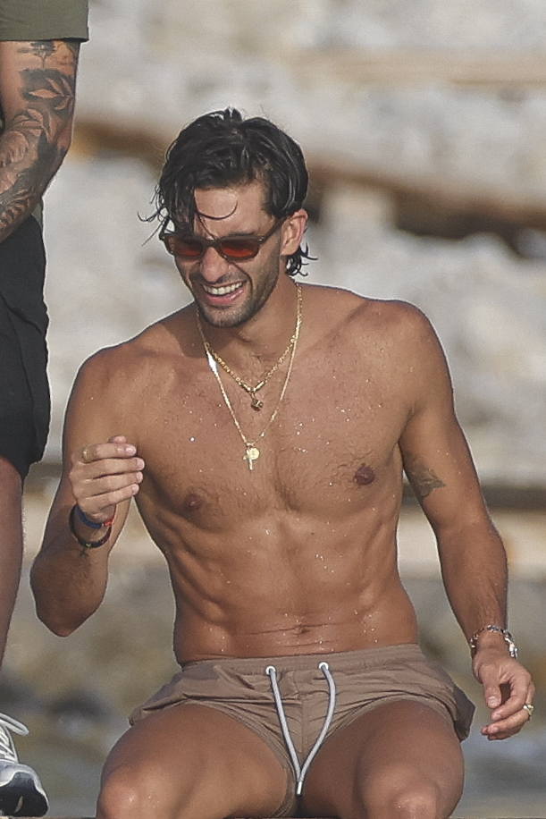 Shirtless man with dark hair and red sunglasses smiling at a rocky beach, wearing gold chains and beige swim shorts.