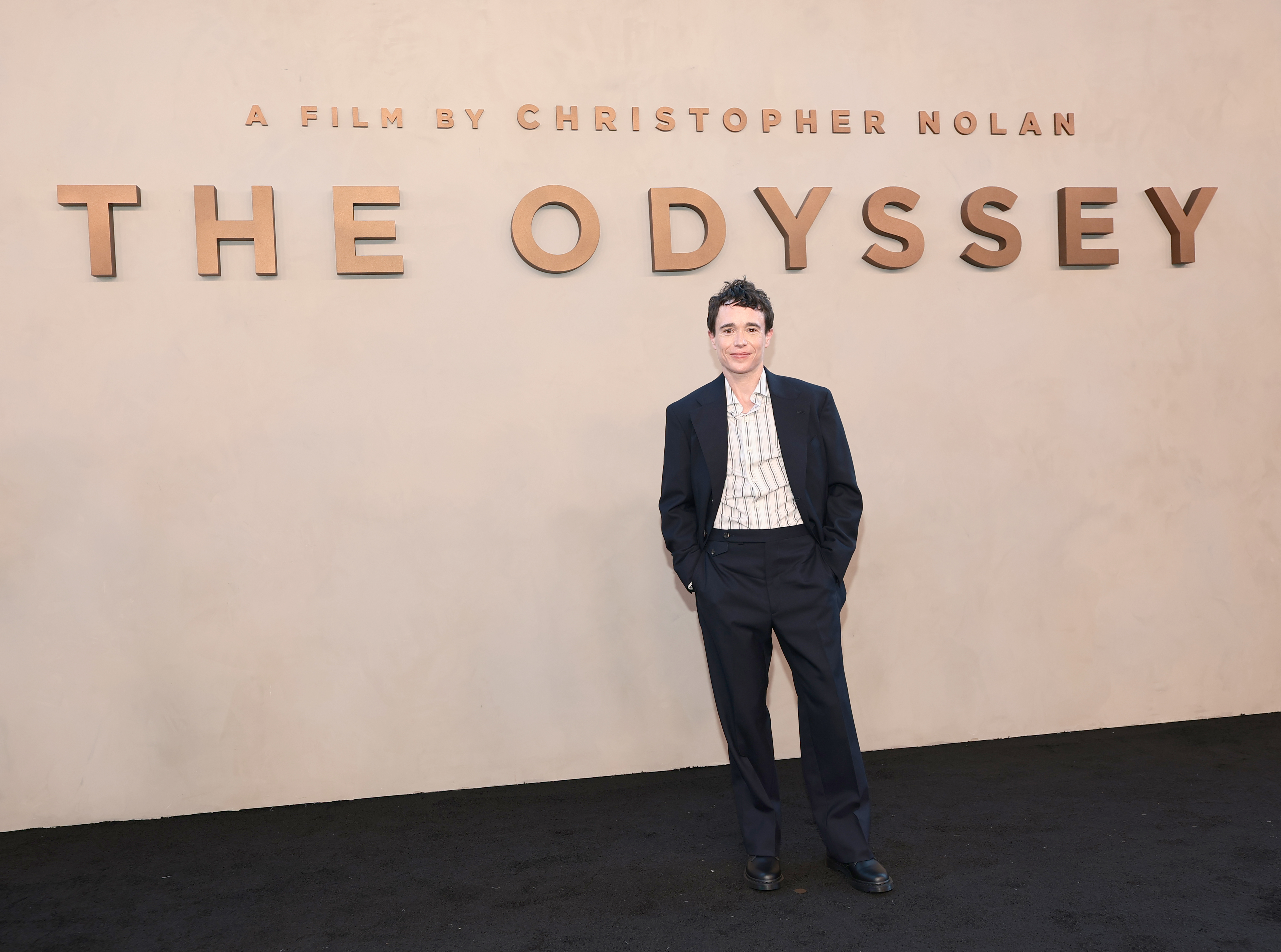 Elliot Page standing in front of a beige wall displaying 'The Odyssey – A Film by Christopher Nolan' at a premiere event.