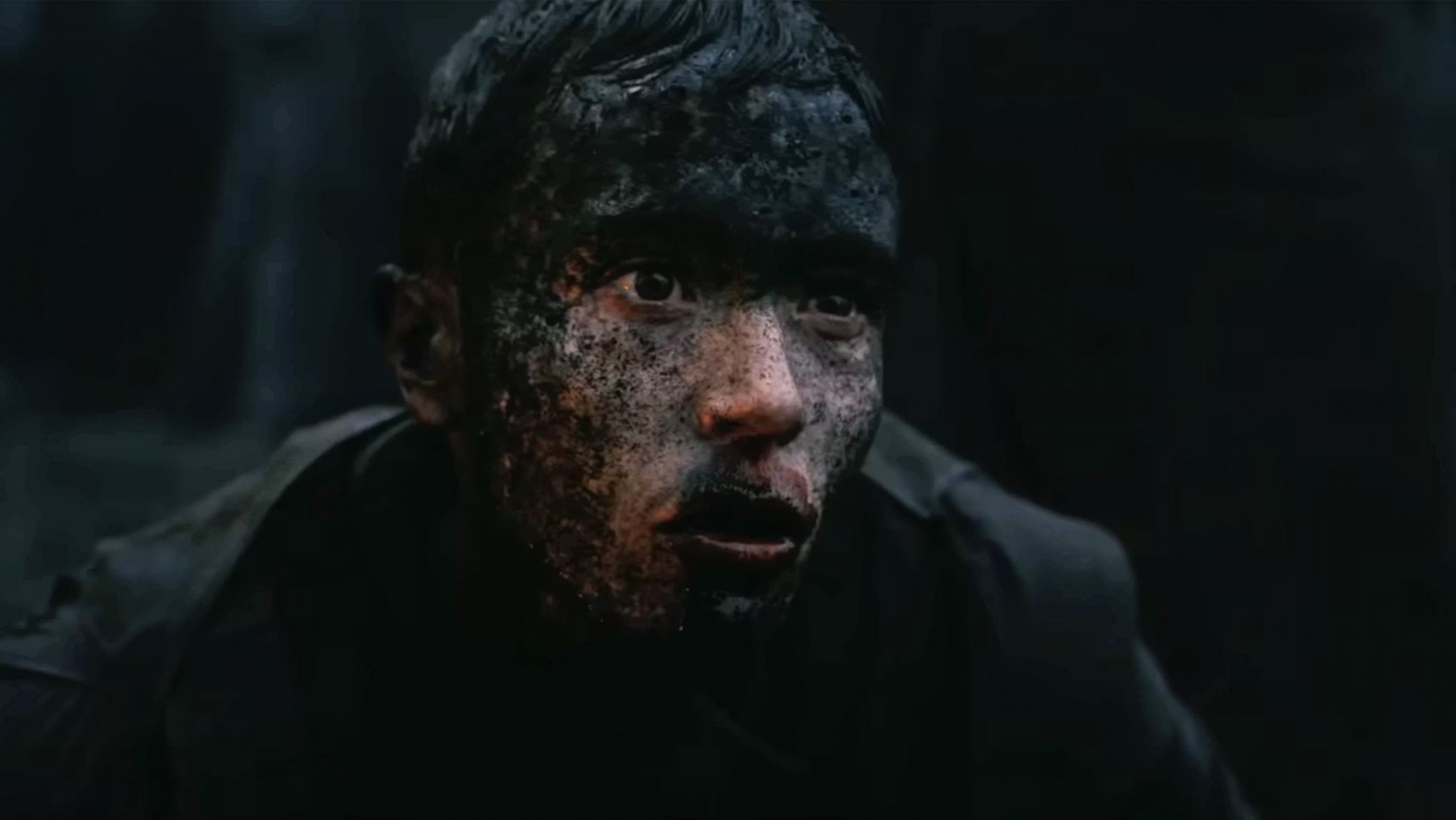 Person with face and hair heavily coated in dark mud staring wide-eyed into camera against a dark background.