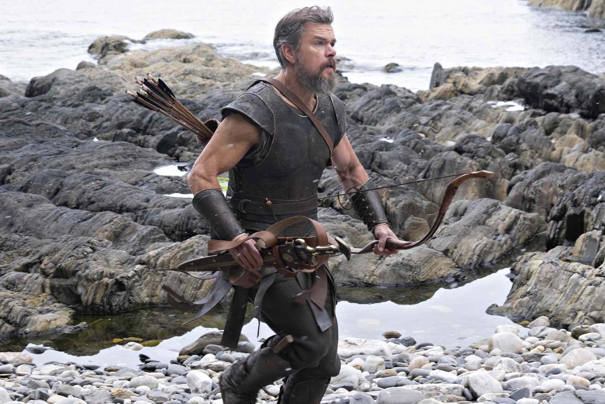 Bearded man in ancient leather armor carrying a bow and quiver of arrows, running across a rocky, pebbled shoreline.