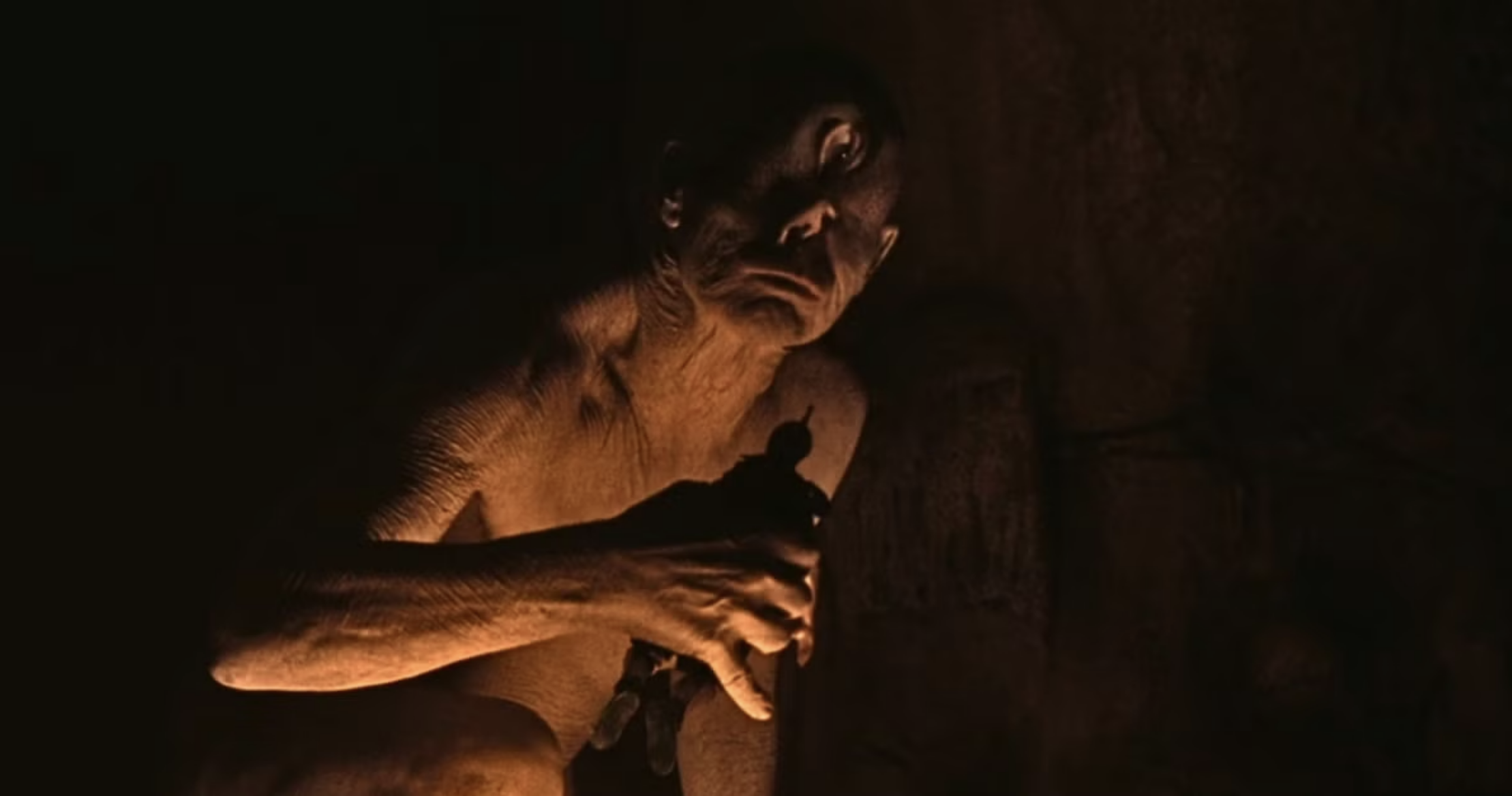 Gaunt, pale humanoid creature crouching in darkness, illuminated by warm amber light, with long limbs and sunken features.